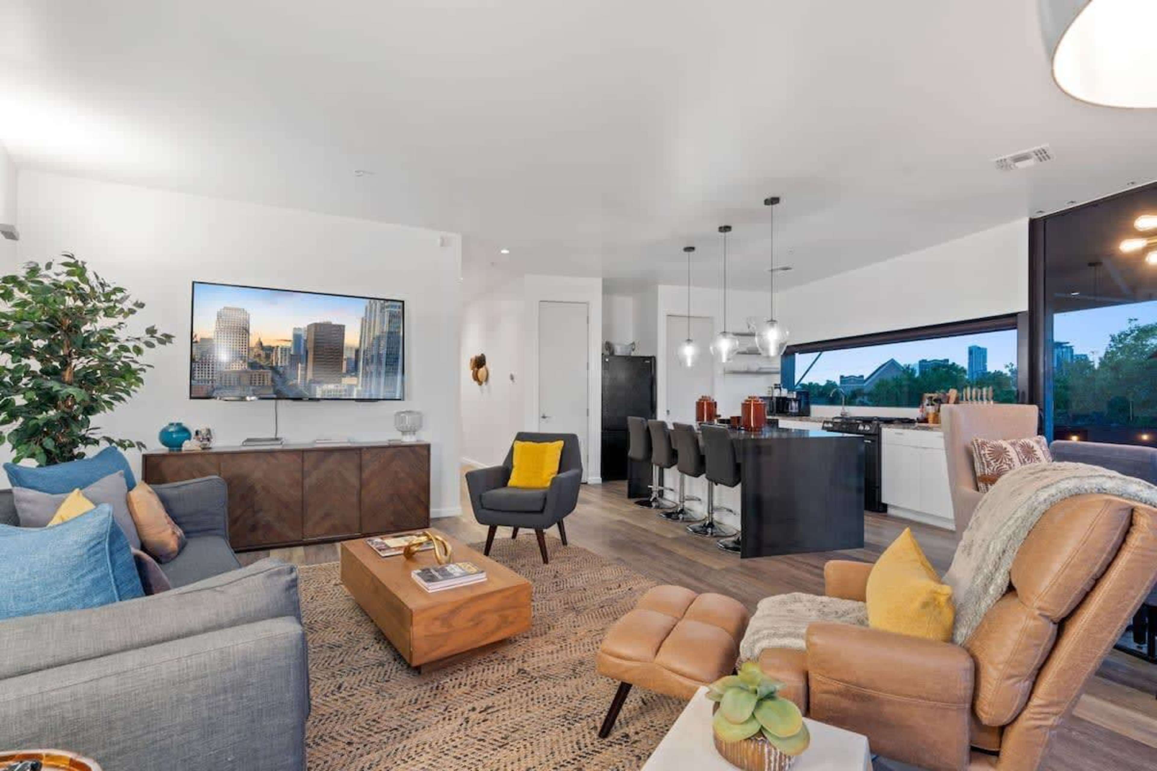 A modern living room with a gray sofa, wooden coffee table, and a large television mounted on the wall, adjacent to a kitchen featuring a breakfast bar and stylish seating.