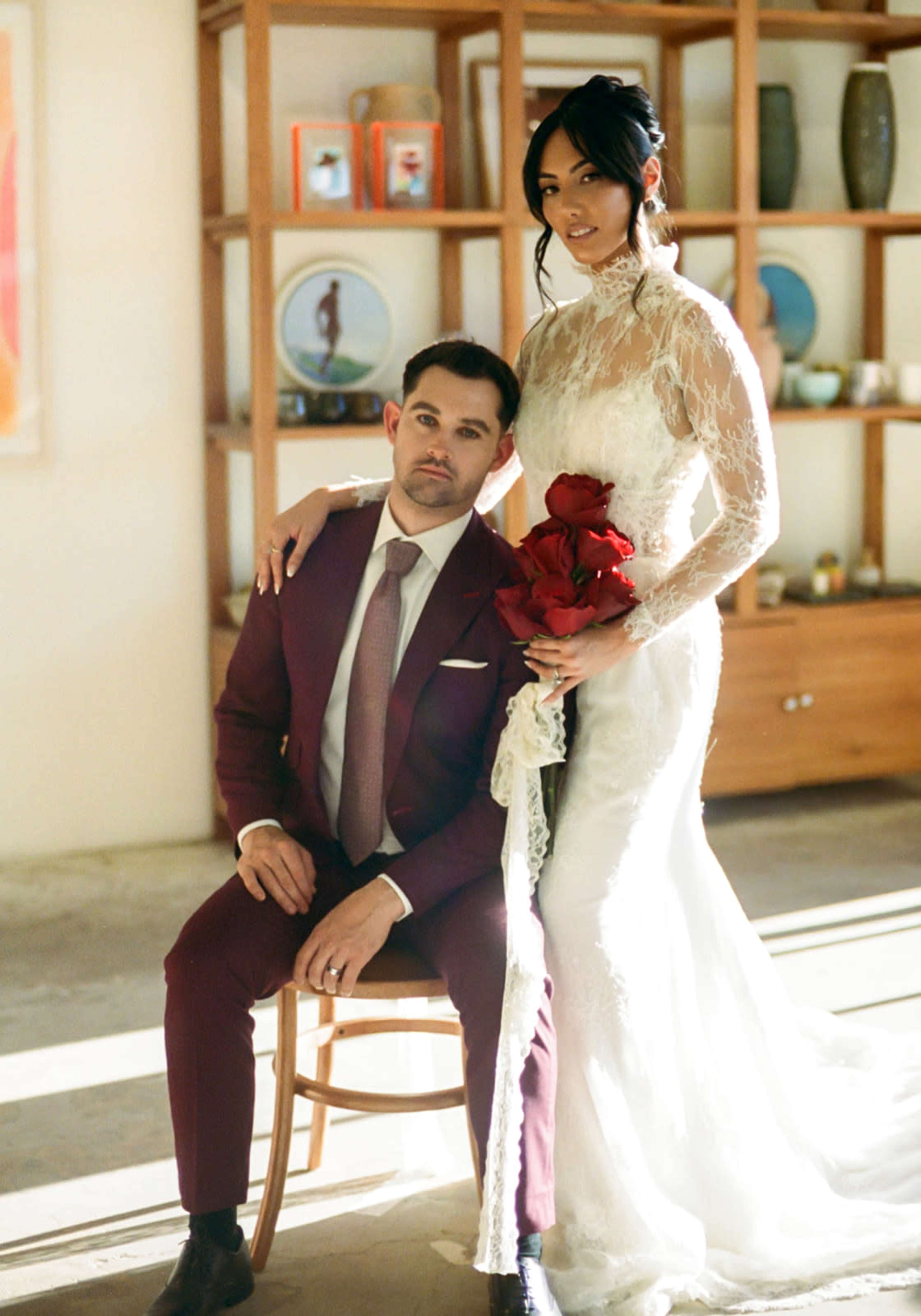 A woman in a white wedding dress holds a bouquet of red roses while seated next to a man in a burgundy suit in a well-lit indoor setting.