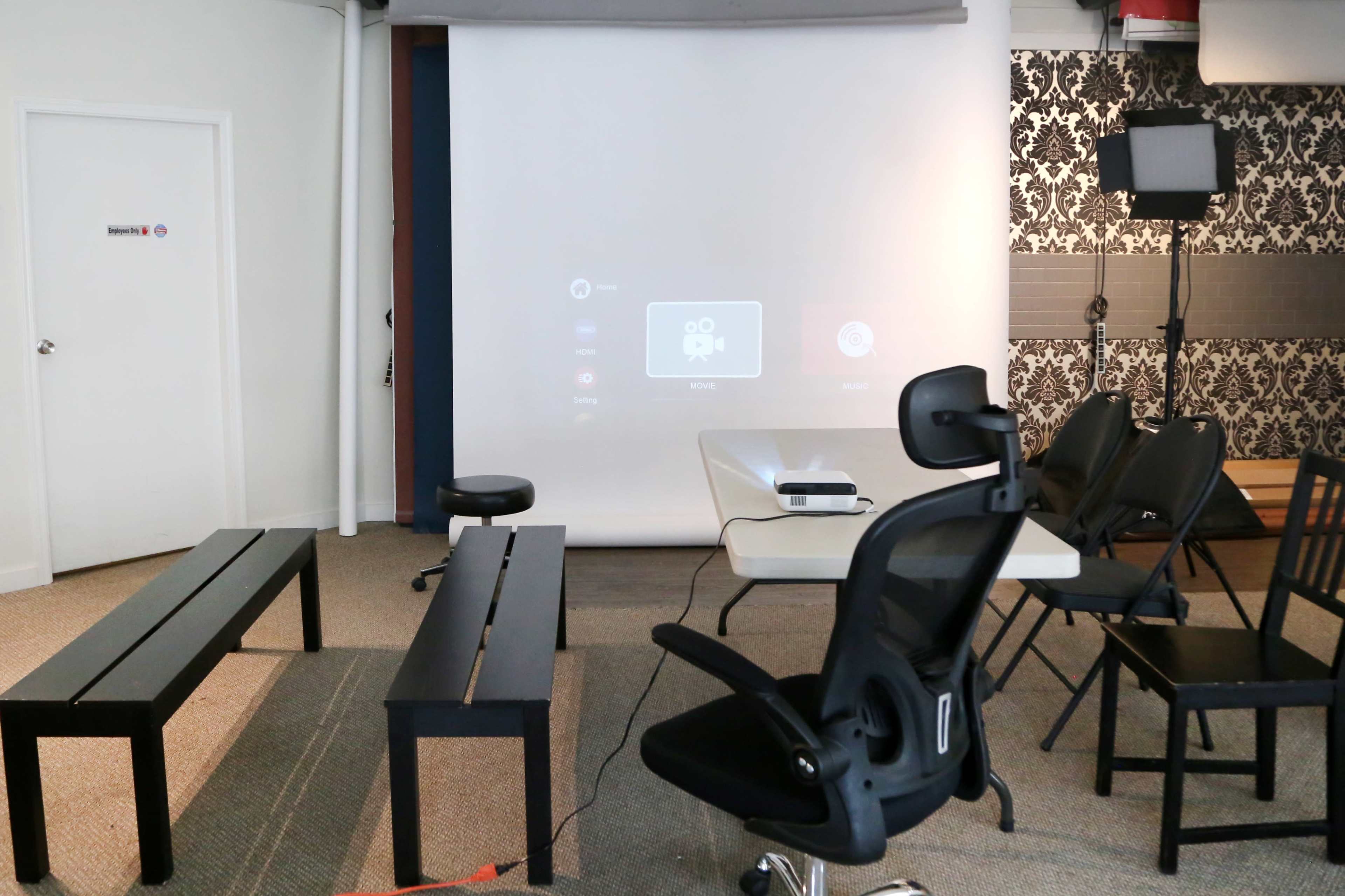 A projector is set up in a room with a white screen, surrounded by chairs and benches arranged for a presentation or meeting.