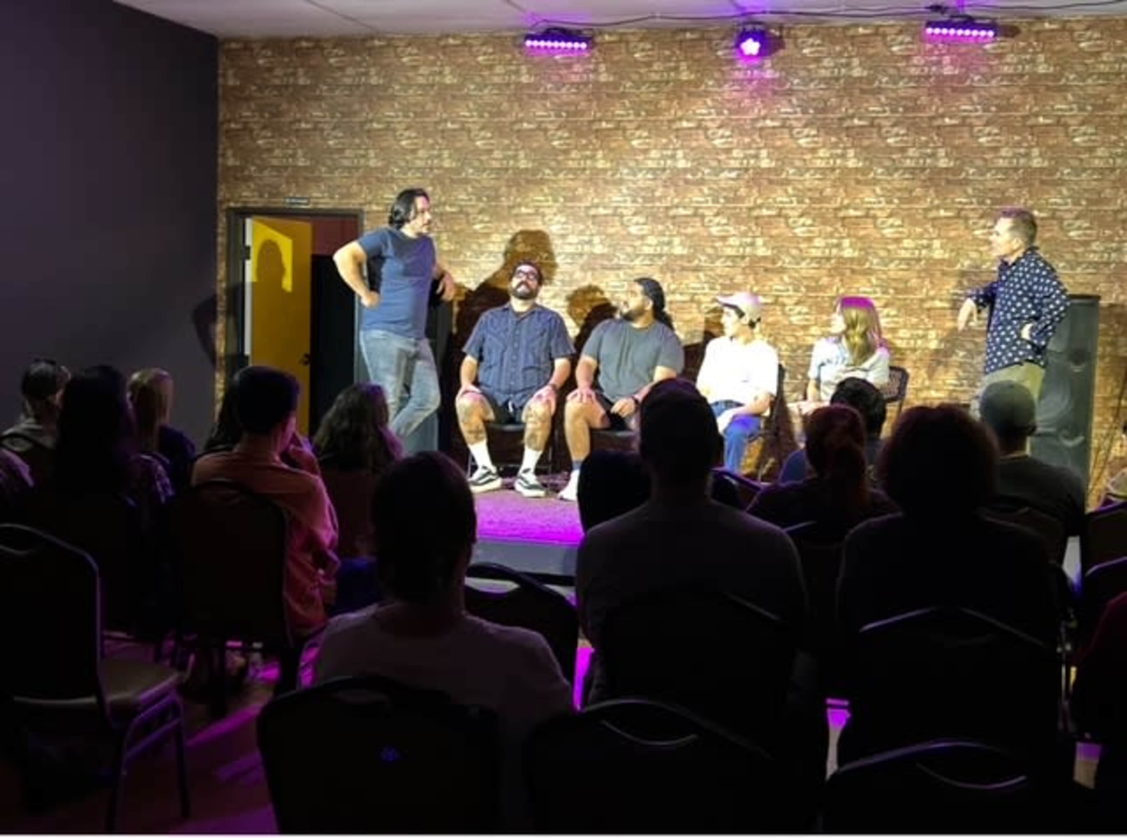 A group of five people sits on stage while an audience watches from their seats in a dimly lit room with a patterned backdrop.