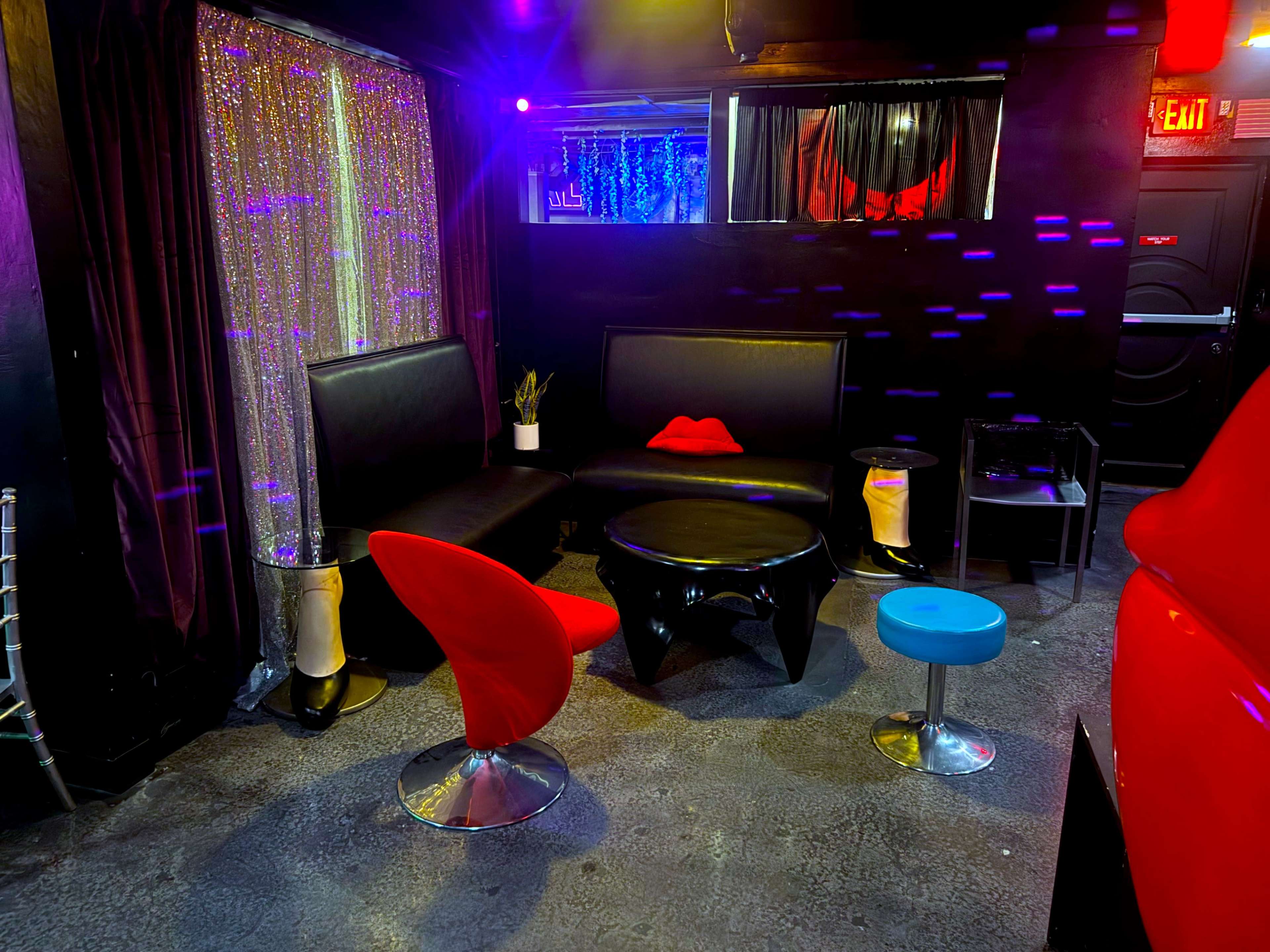 The image shows a dimly lit lounge area with colorful lighting, featuring modern furniture including black and red chairs, a circular black table, and a decorative curtain in the background.