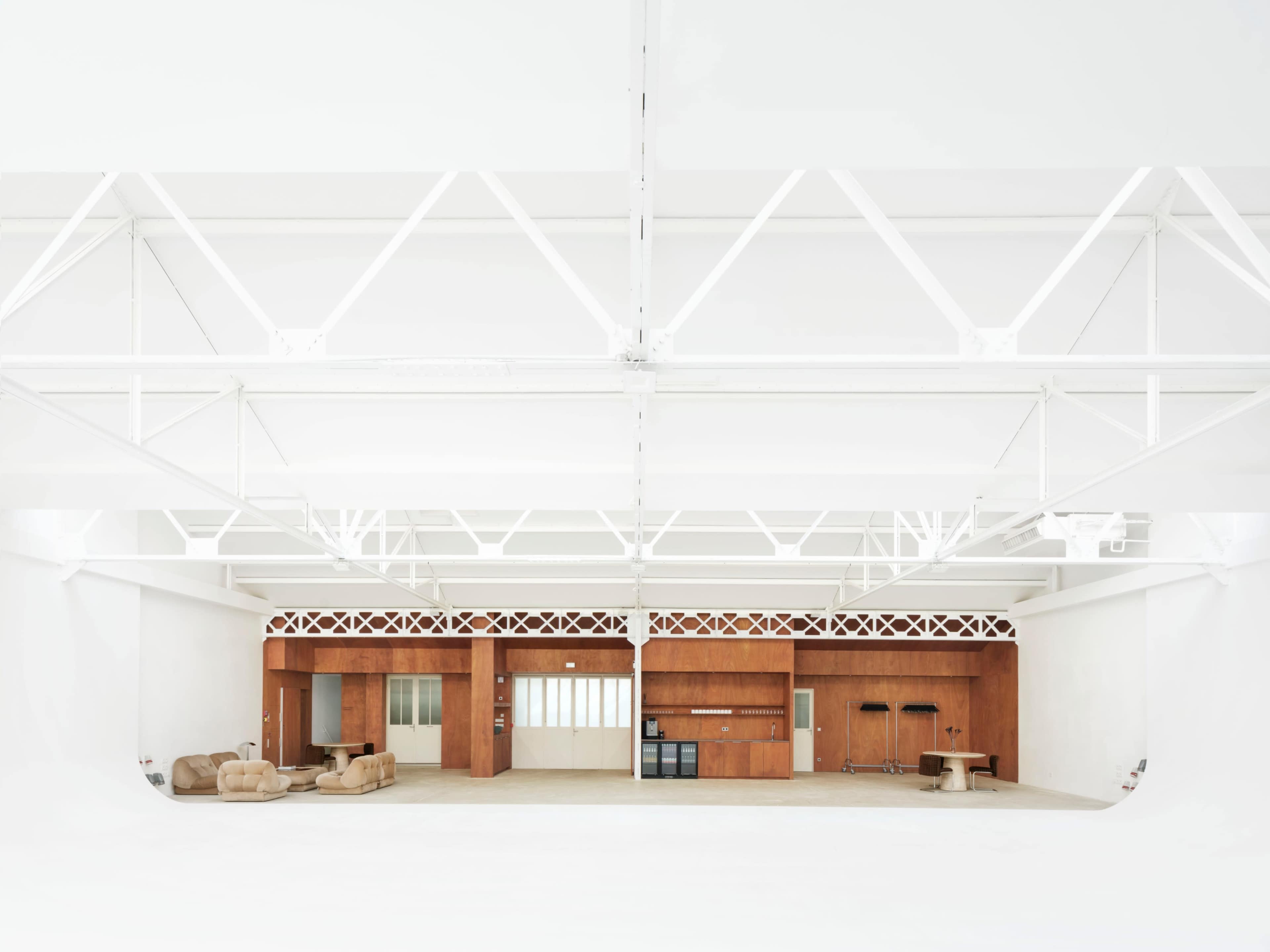 The image shows a spacious, minimalist interior featuring wooden cabinetry, a couple of beige sofas, and a high, airy ceiling supported by white beams.