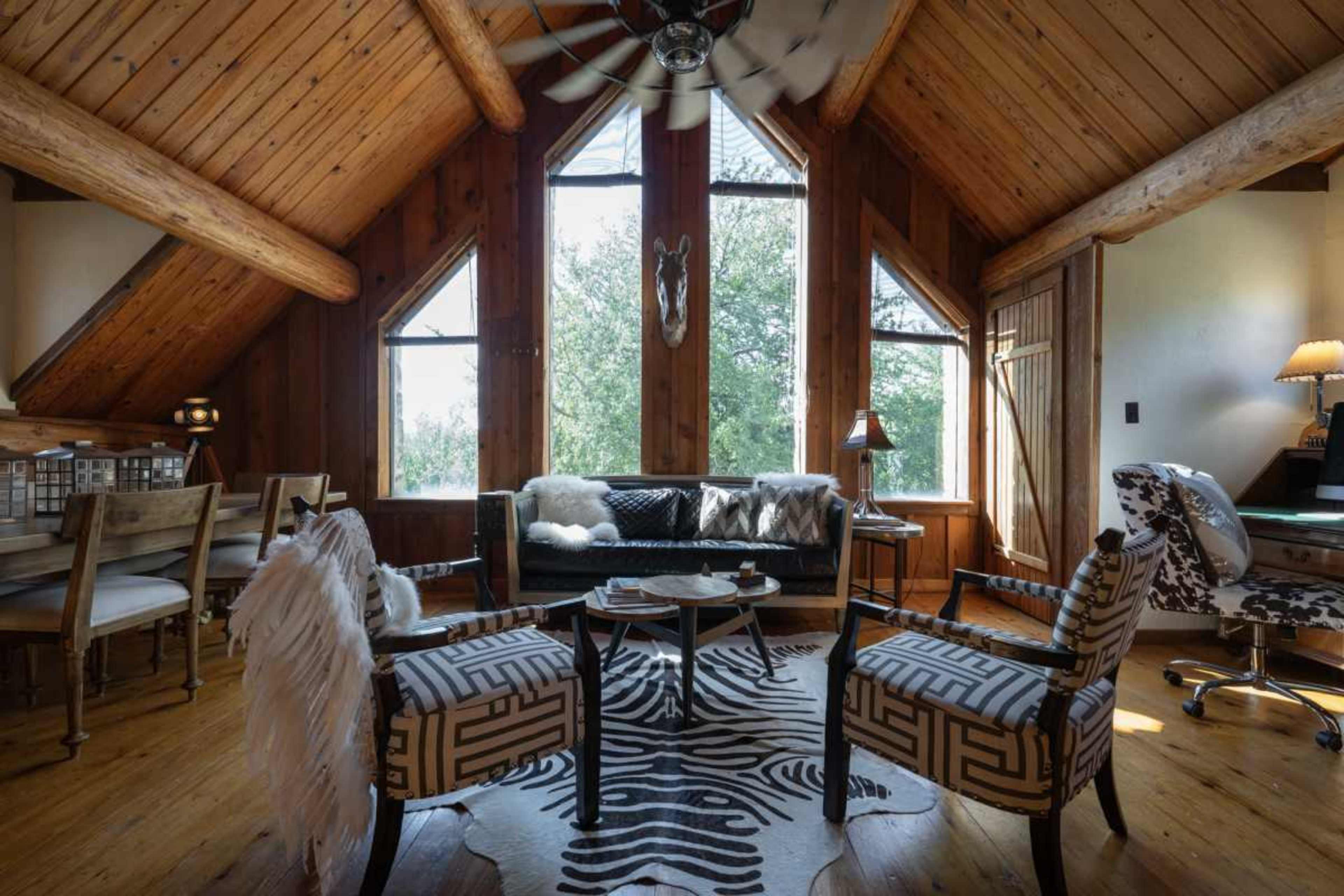 The image shows a cozy, wooden living room with high ceilings, large triangular windows, and a mix of modern and rustic furnishings.