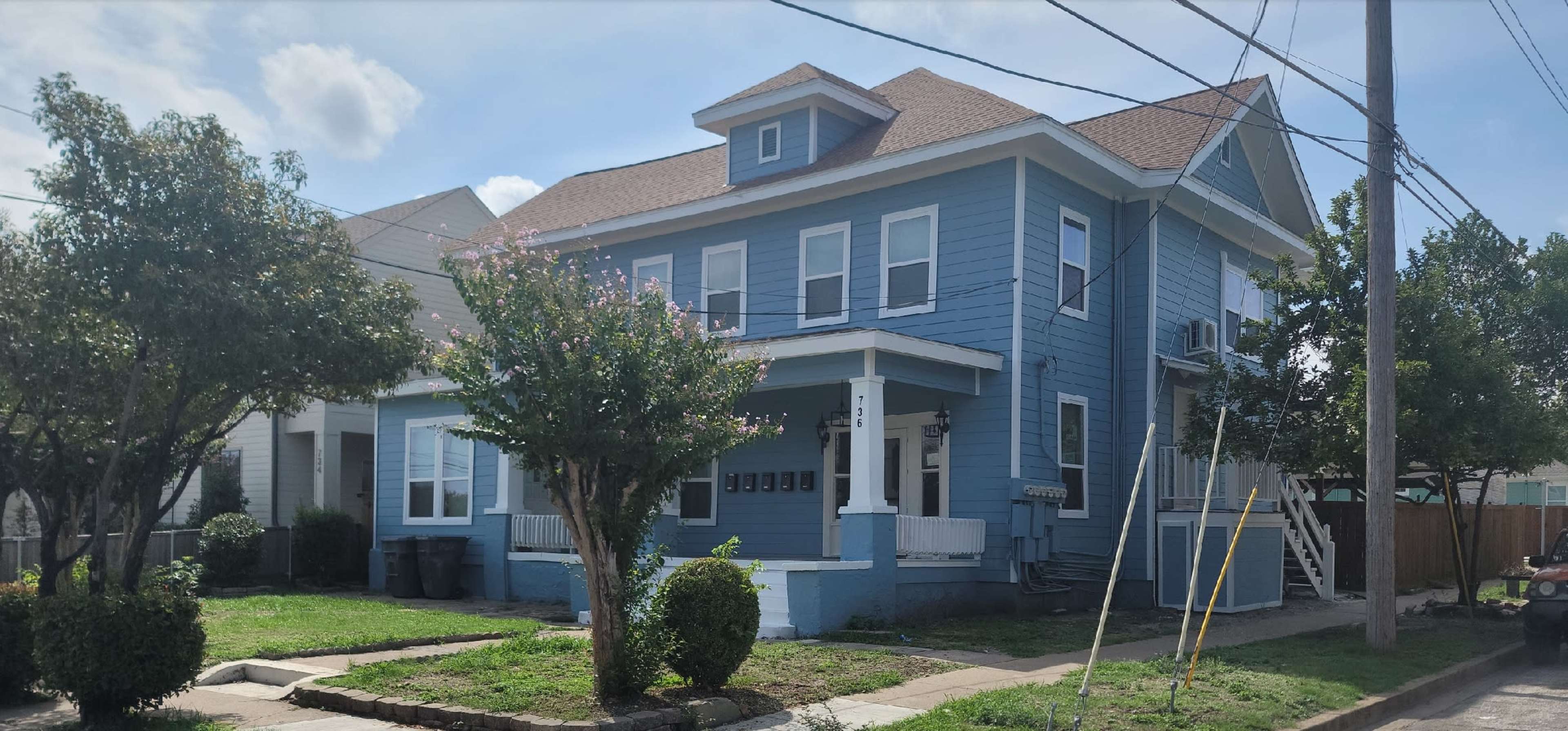 A two-story blue house with a porch and trees in front, situated on a residential street.