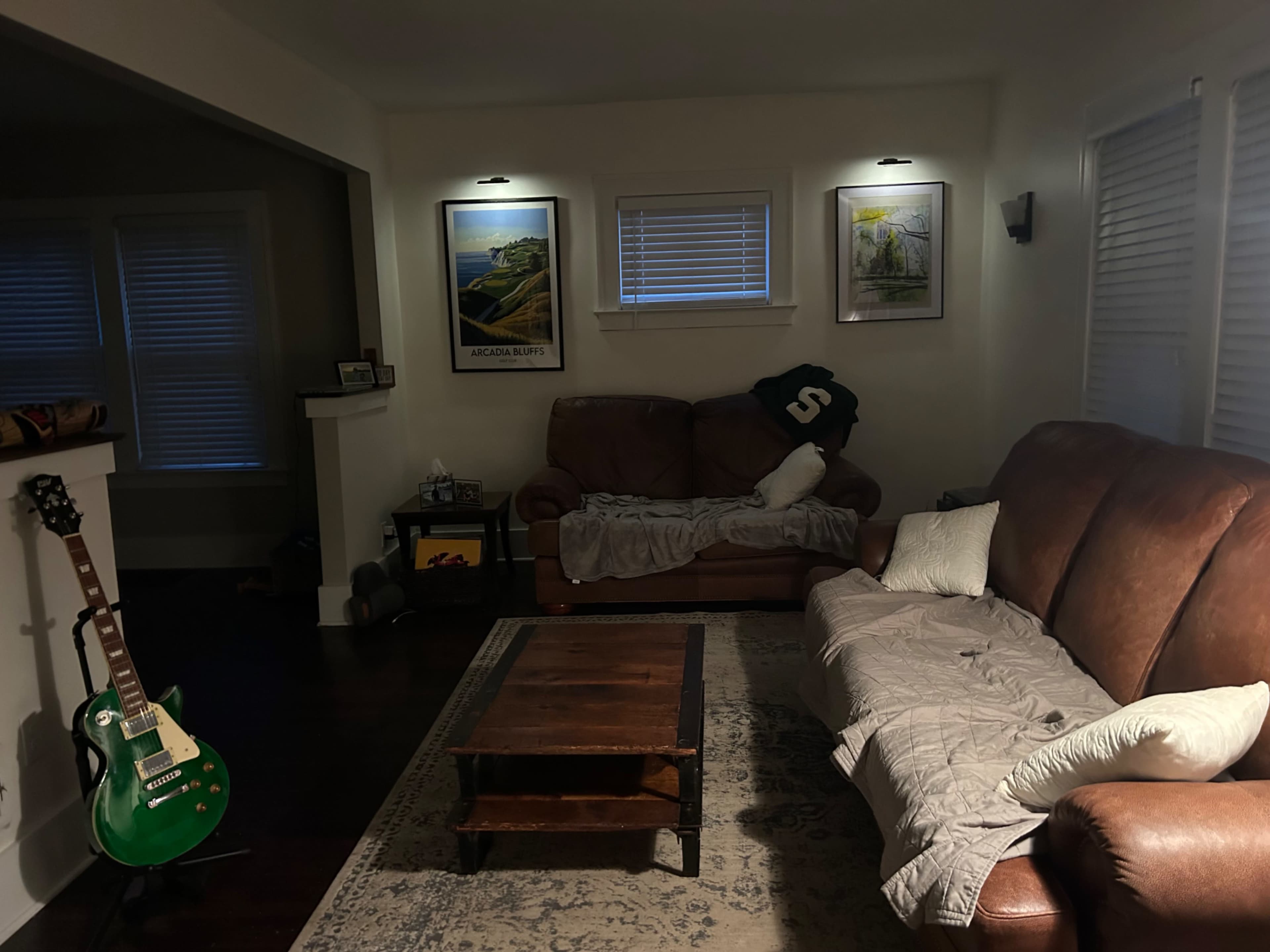 The living room features a brown leather couch, a coffee table, and a green guitar resting against the wall, with framed artwork and lamps illuminating the space.