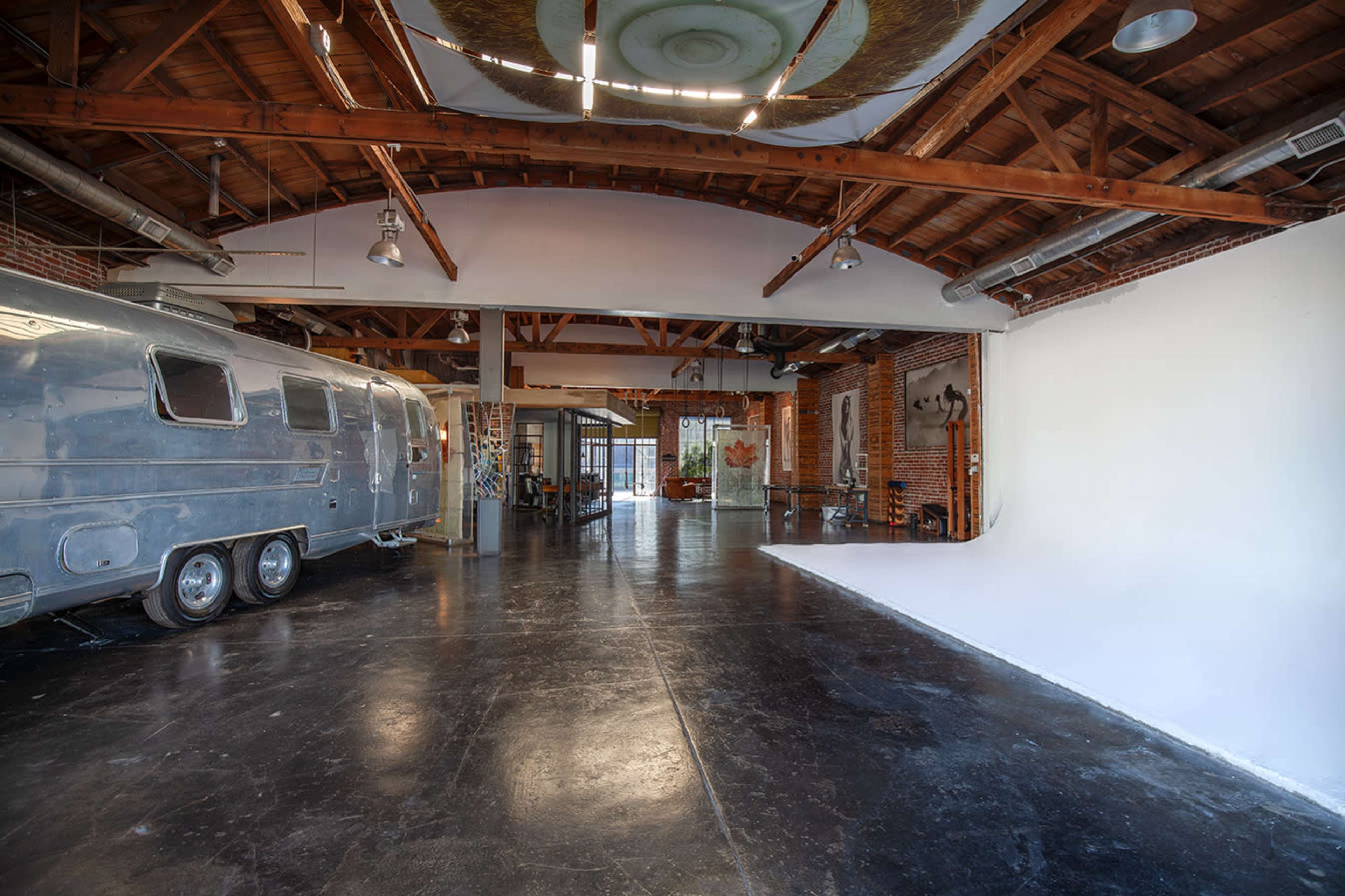 A spacious interior with high wooden ceilings, featuring a vintage trailer on one side and a blank white wall on the other.