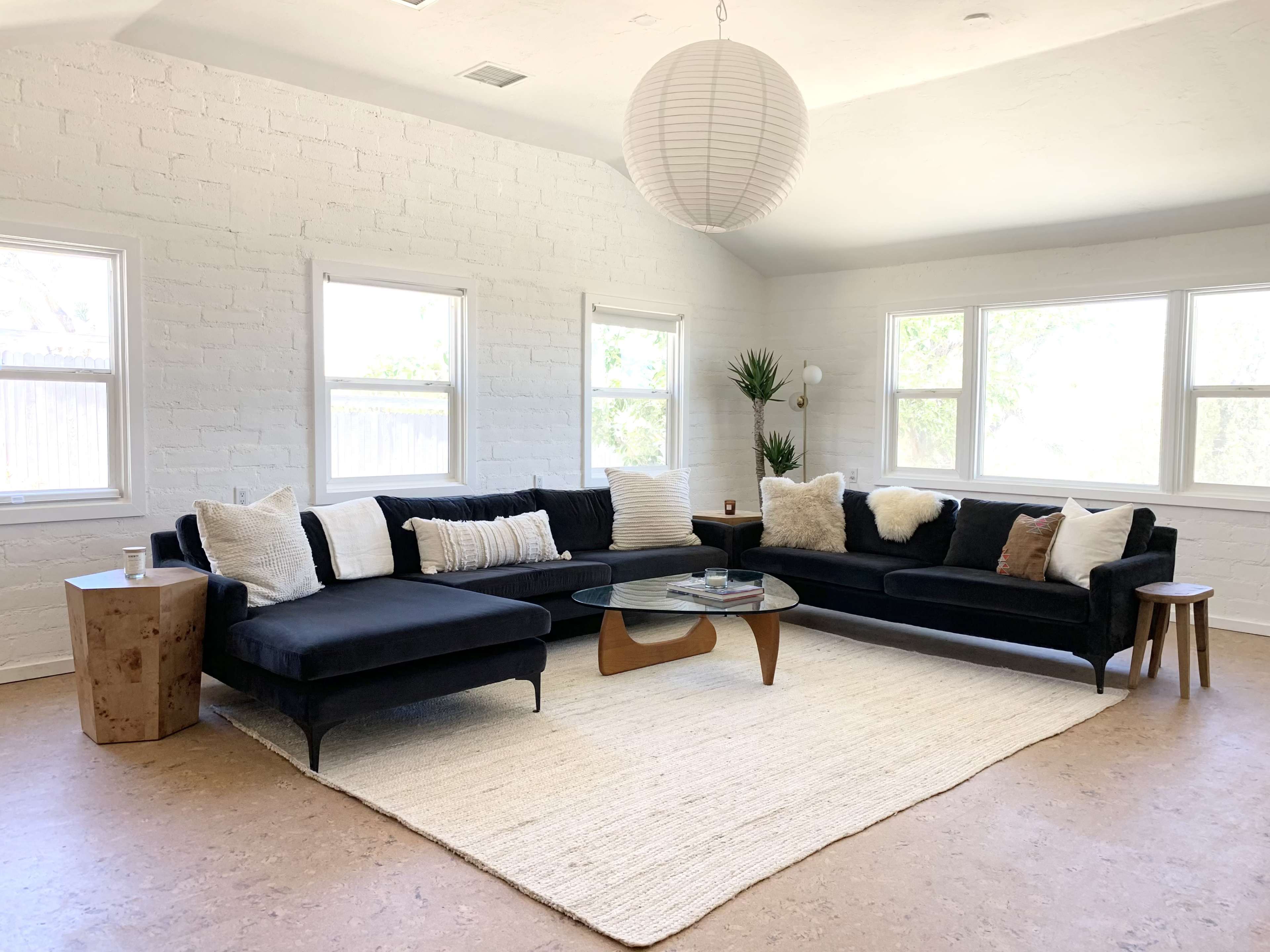 A spacious living room features a large black sectional sofa, a round glass coffee table, and a light-colored area rug, surrounded by white walls and multiple windows.