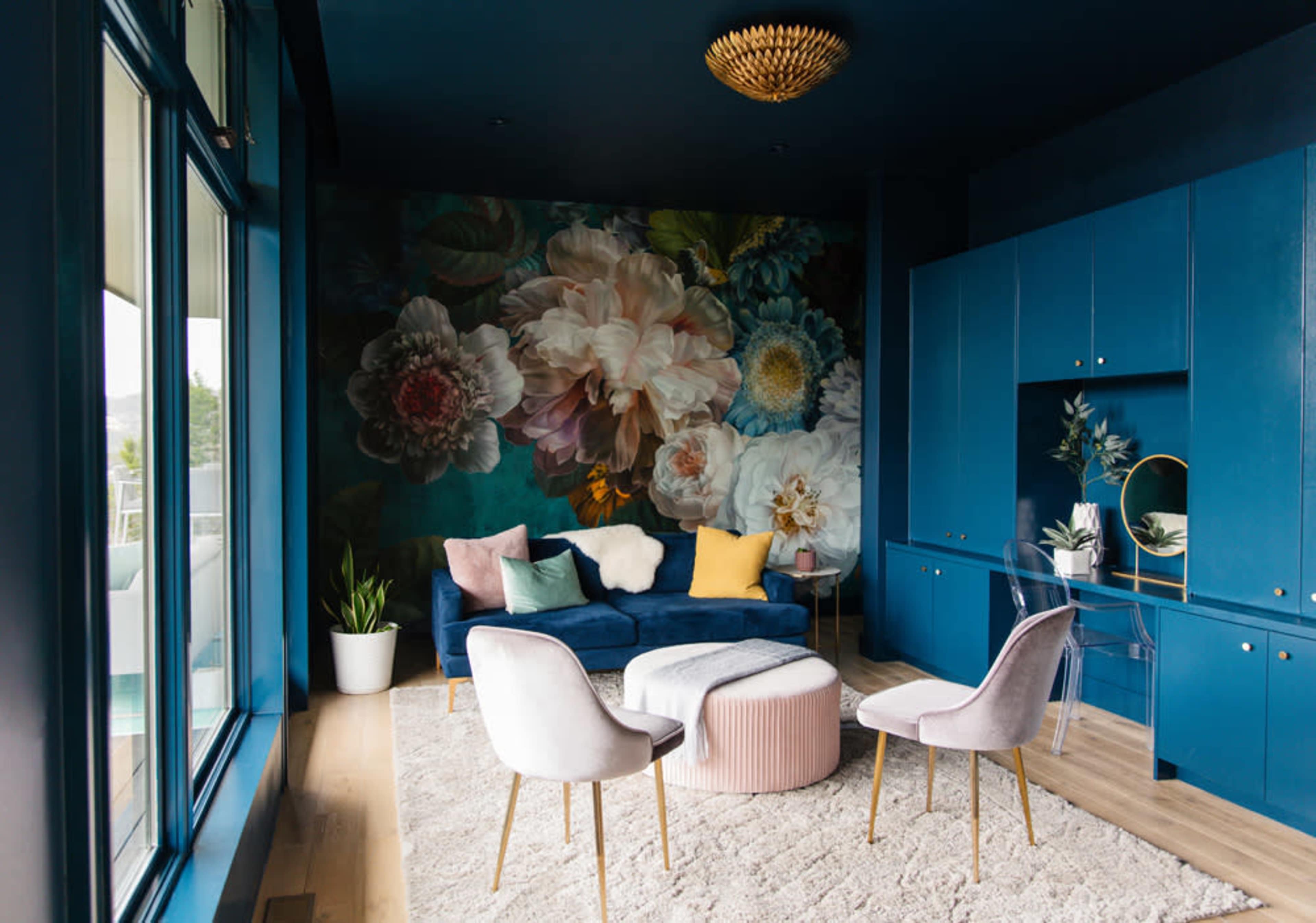 A modern living room features a floral mural, a dark blue sofa, a circular coffee table, and two white chairs against a backdrop of large windows.