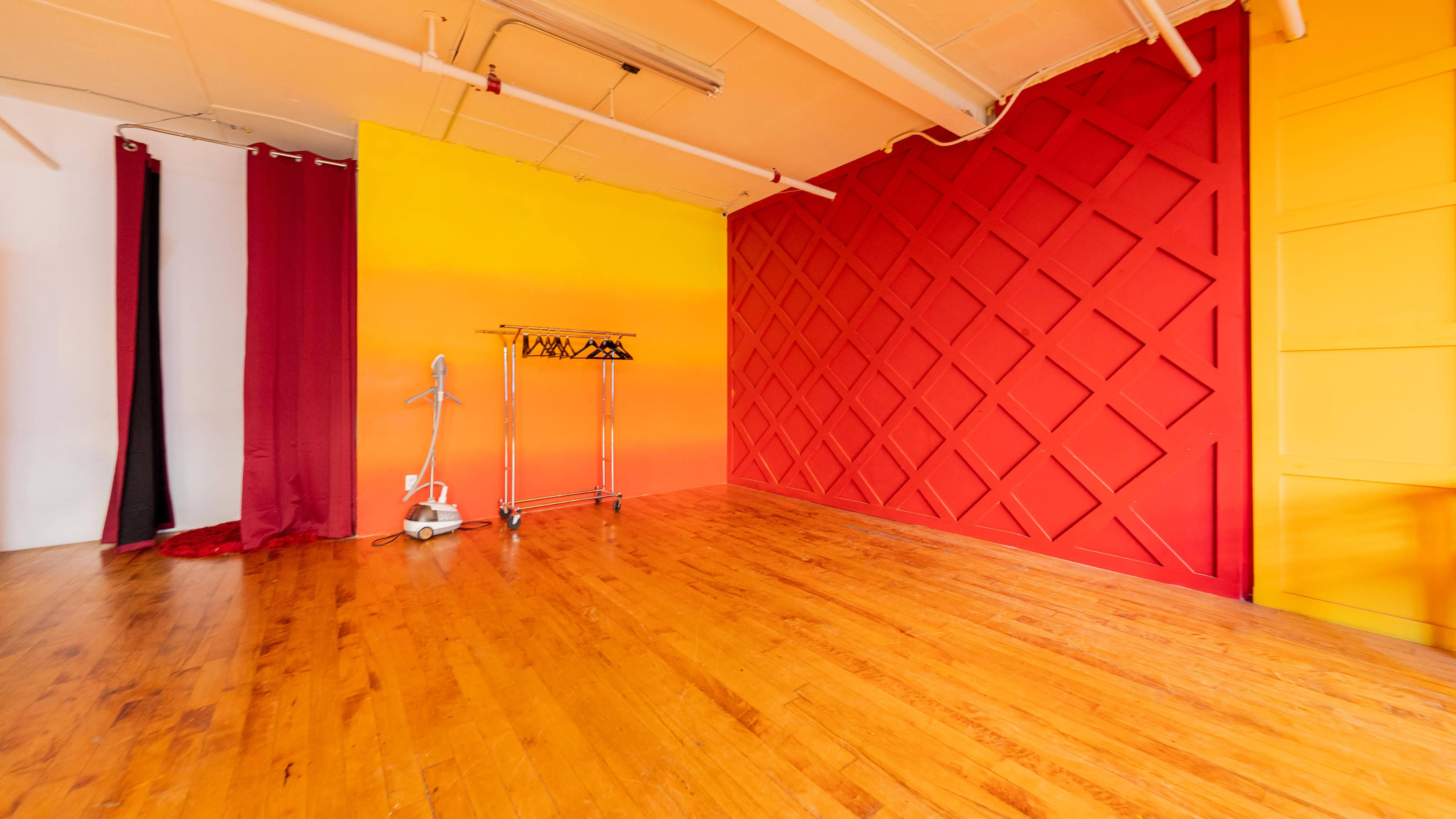 An empty room with wooden flooring, featuring a red and yellow accent wall, two long curtains, and a clothing rack on wheels.