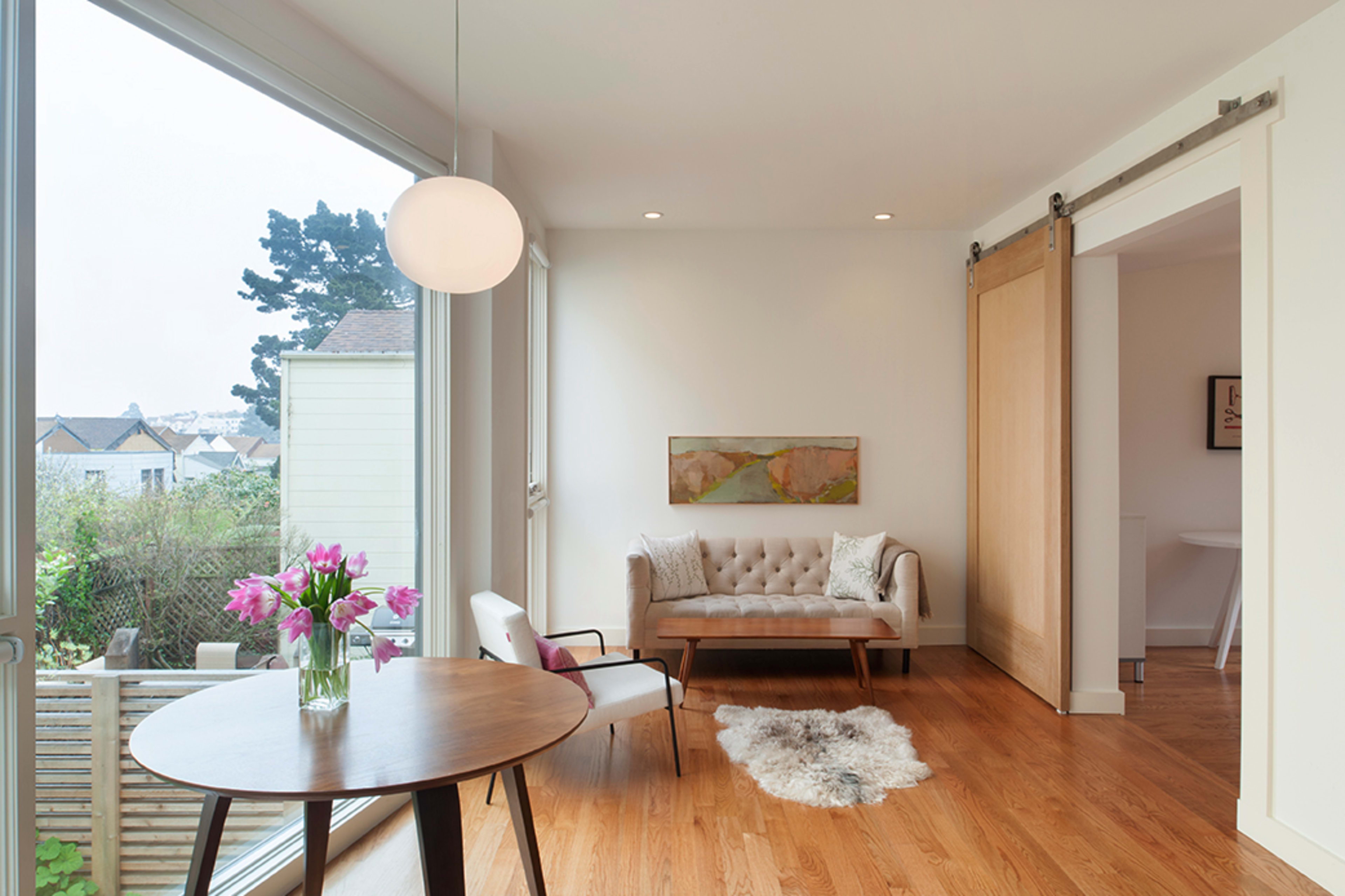 A cozy living space features a light-colored sofa, a round table with a vase of flowers, and large windows allowing natural light to enter.