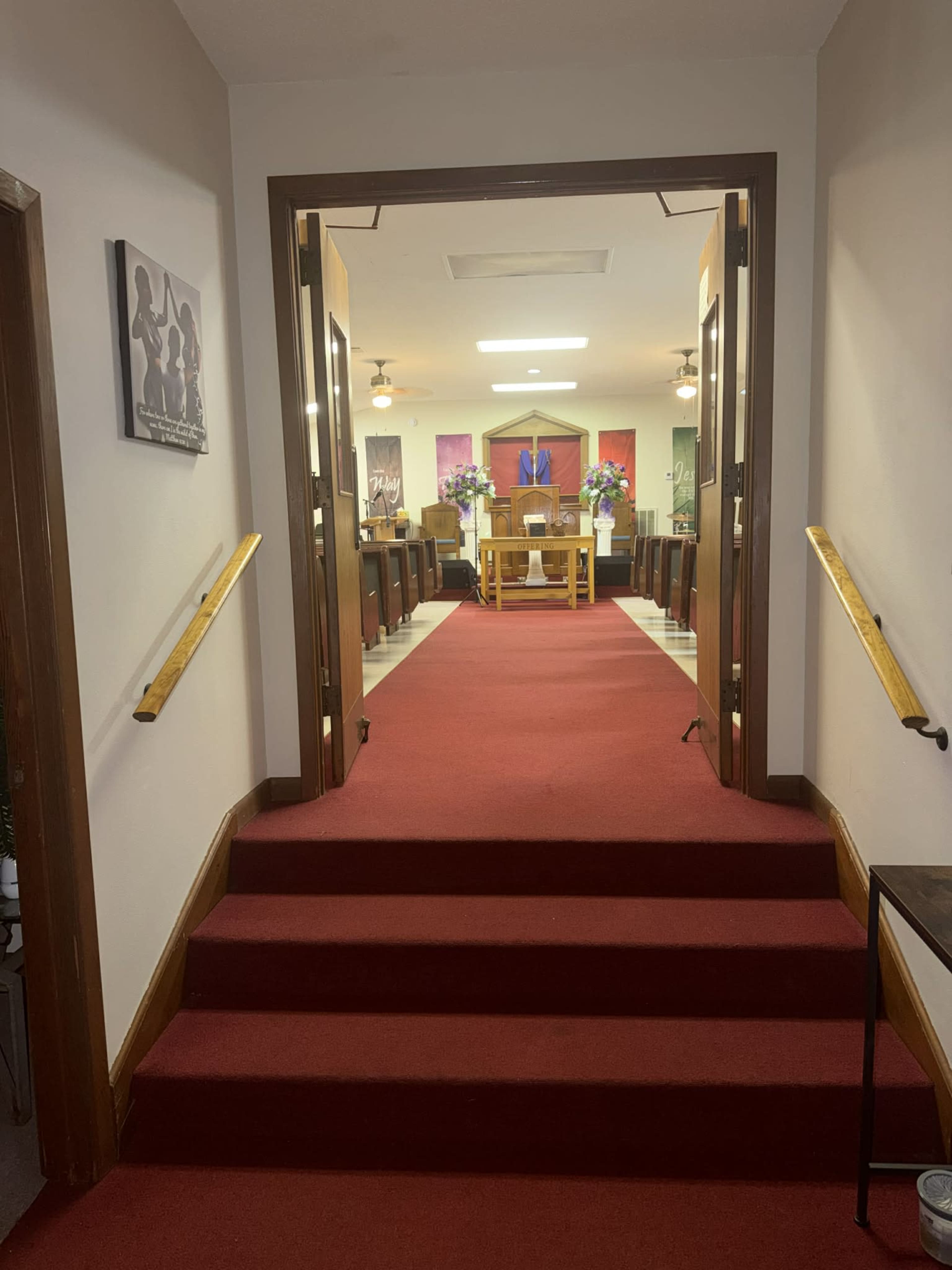 The image shows a view down a carpeted hallway leading to a small chapel or meeting room, with wooden double doors open to reveal a center altar surrounded by flower arrangements.