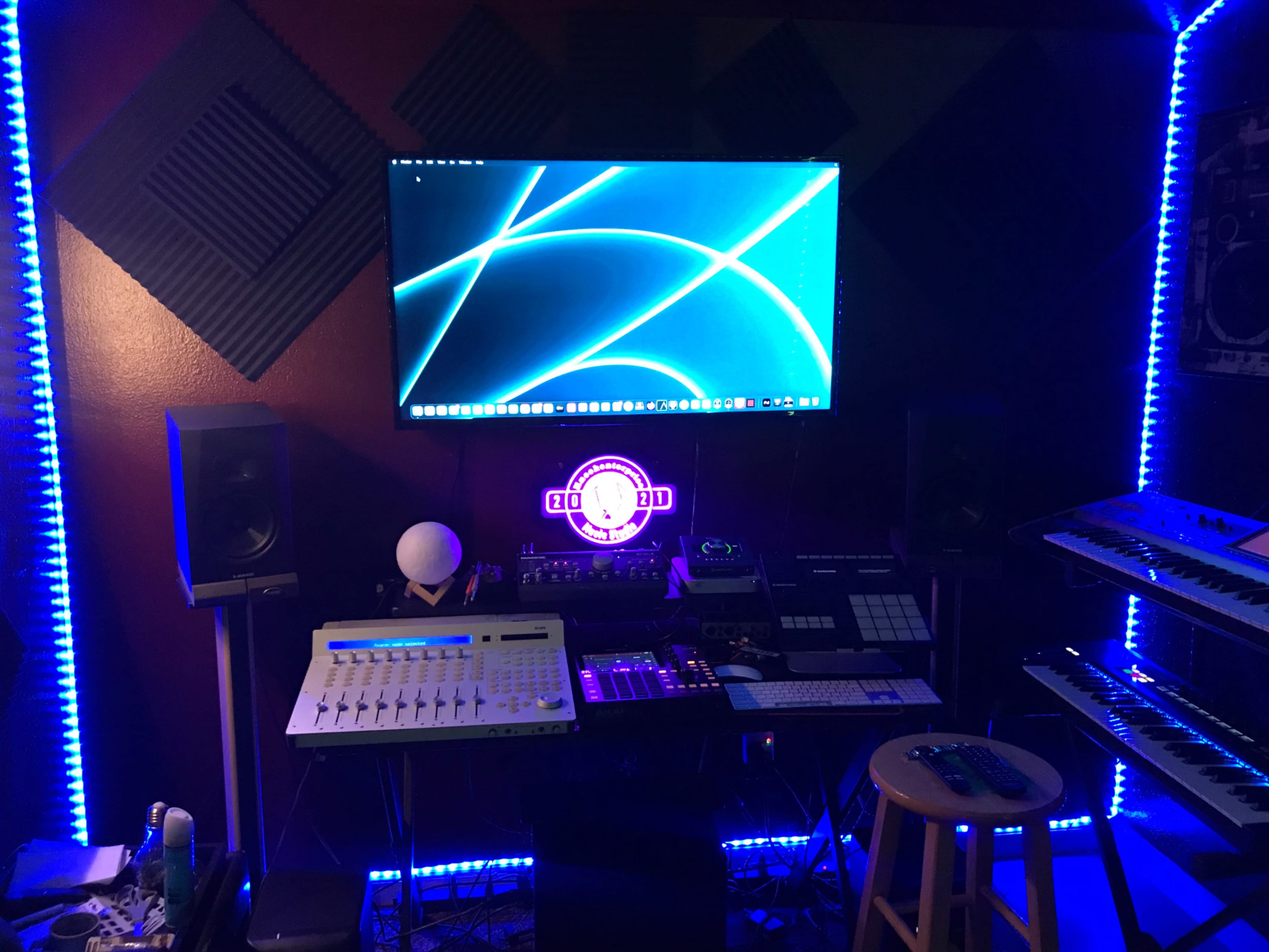A music production studio features a computer screen displaying blue graphics, various electronic instruments, and ambient lighting.