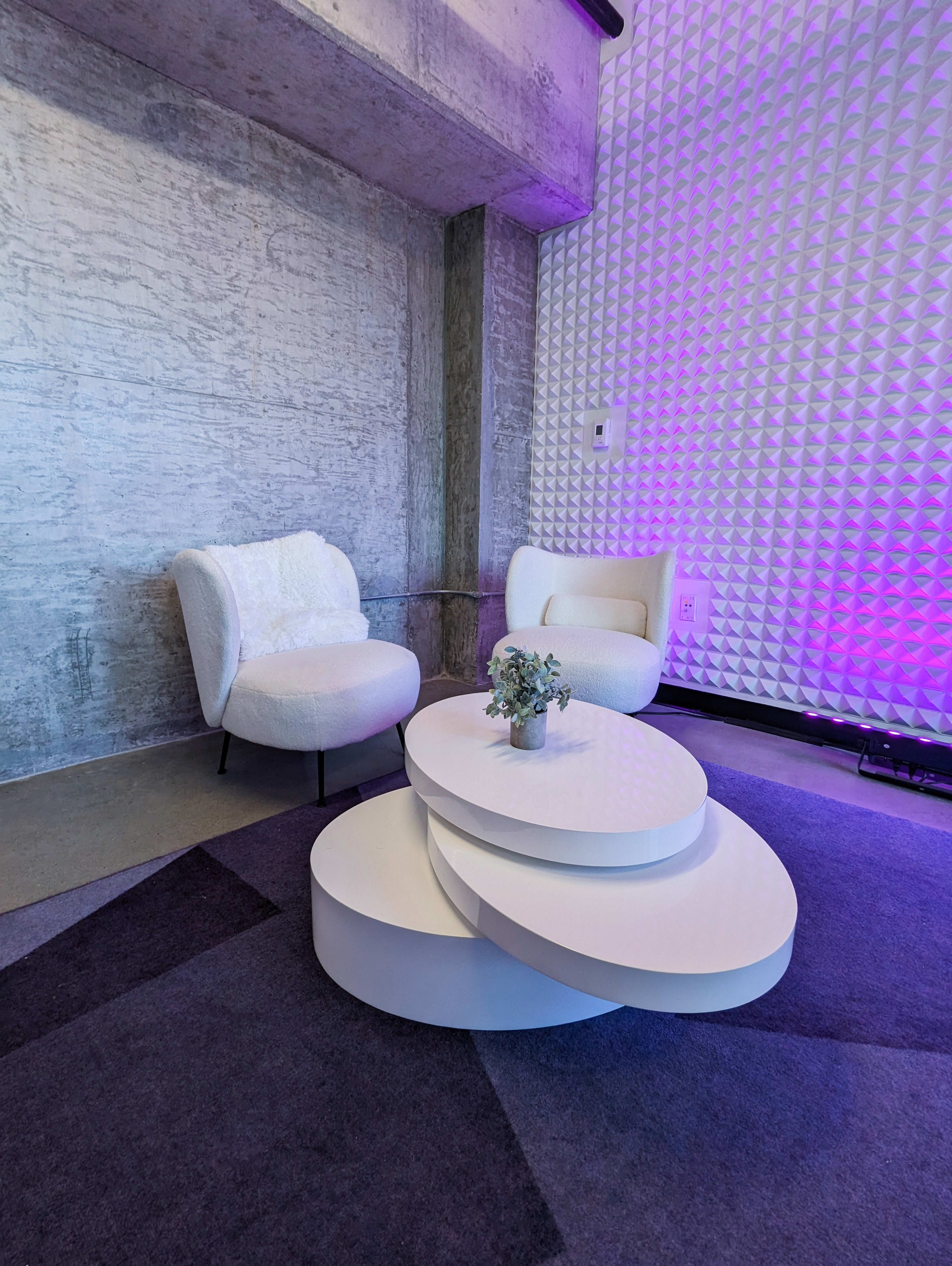 The image shows a modern seating area with two white chairs and a tiered circular coffee table on a patterned rug, set against a textured wall.