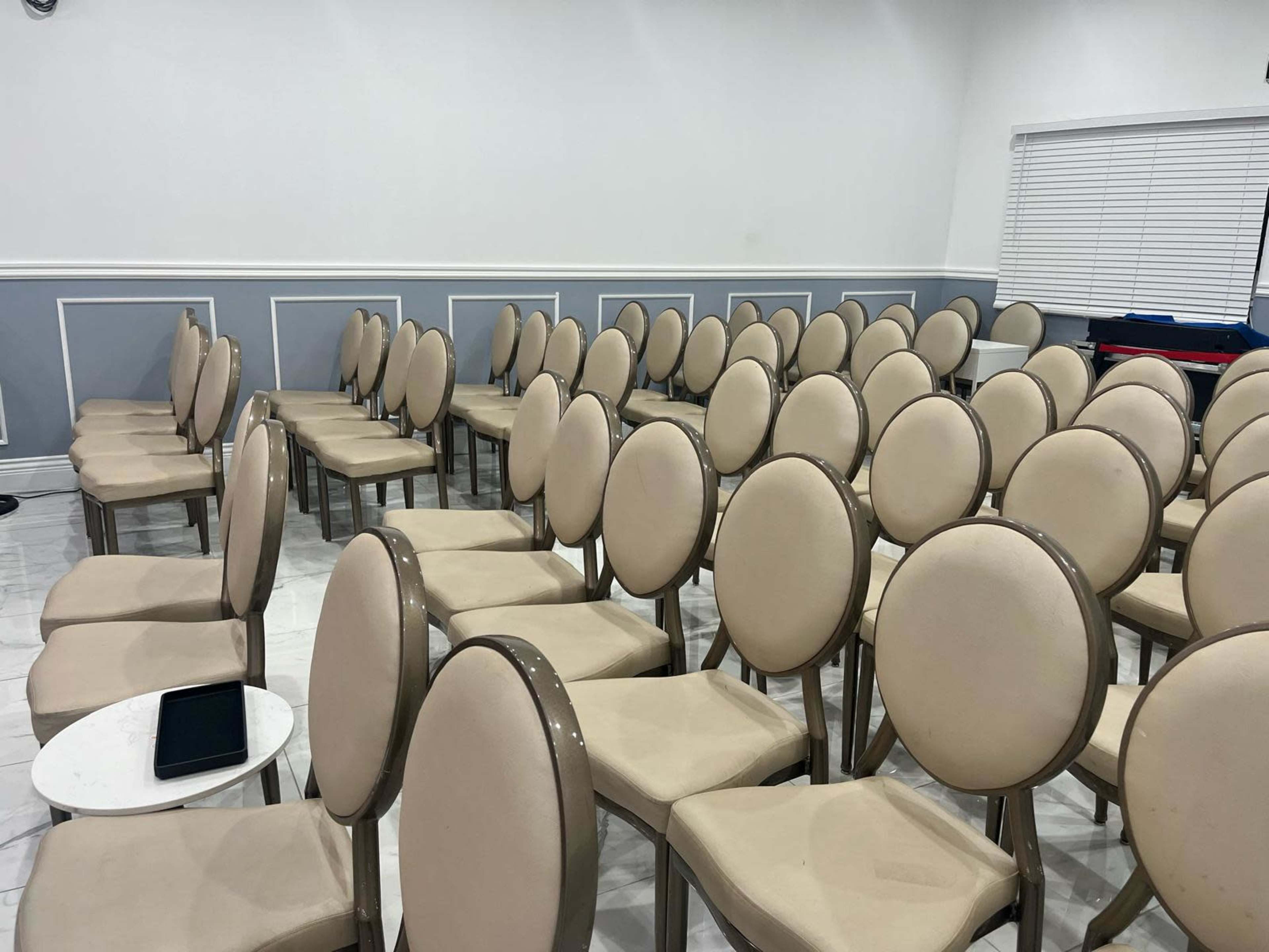A room with rows of beige chairs arranged in a seating layout, with a small table and an electronic device placed nearby.