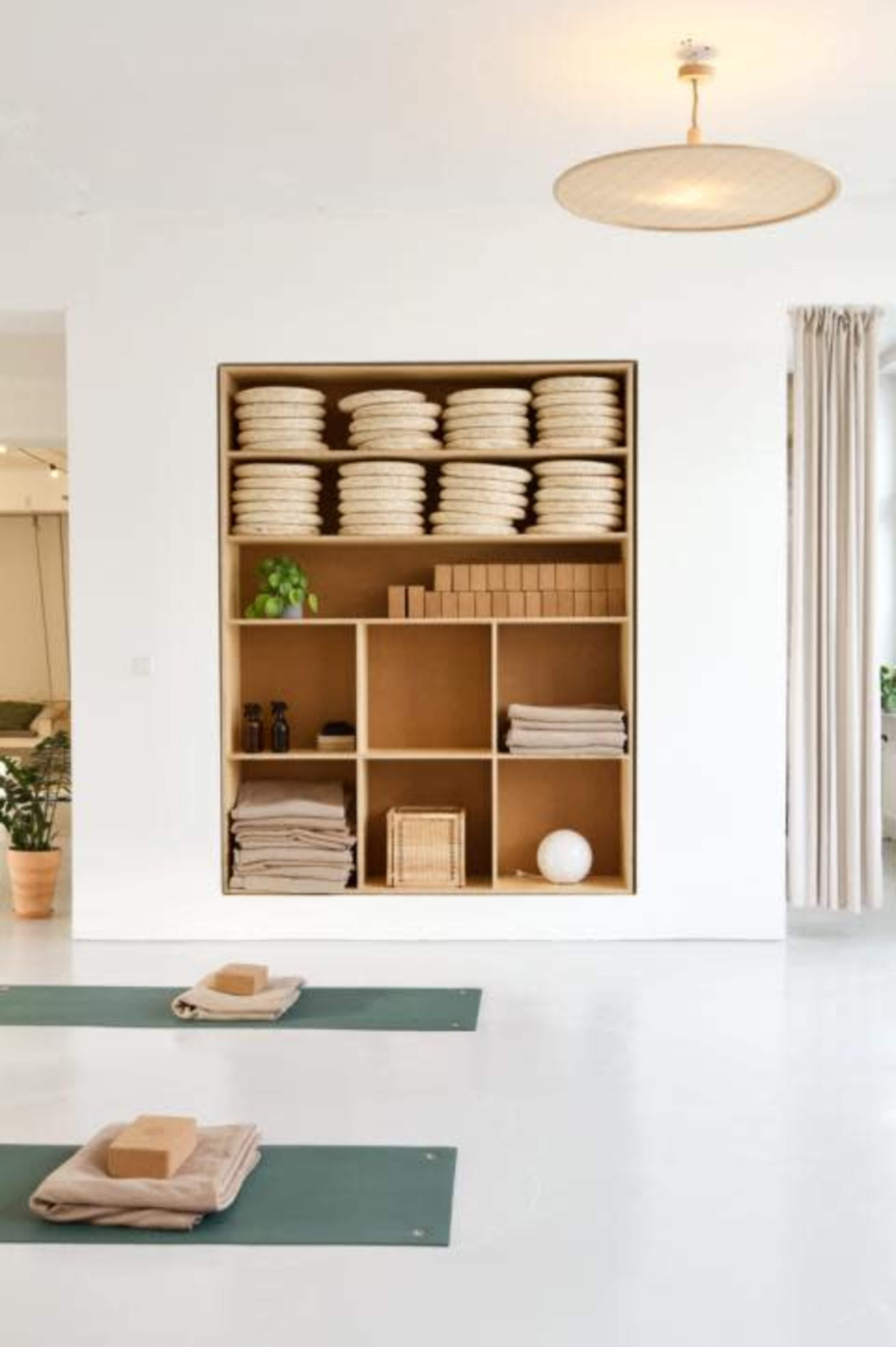A clean, minimalist yoga studio features neatly arranged shelves of yoga mats, blocks, and towels against a bright white wall.