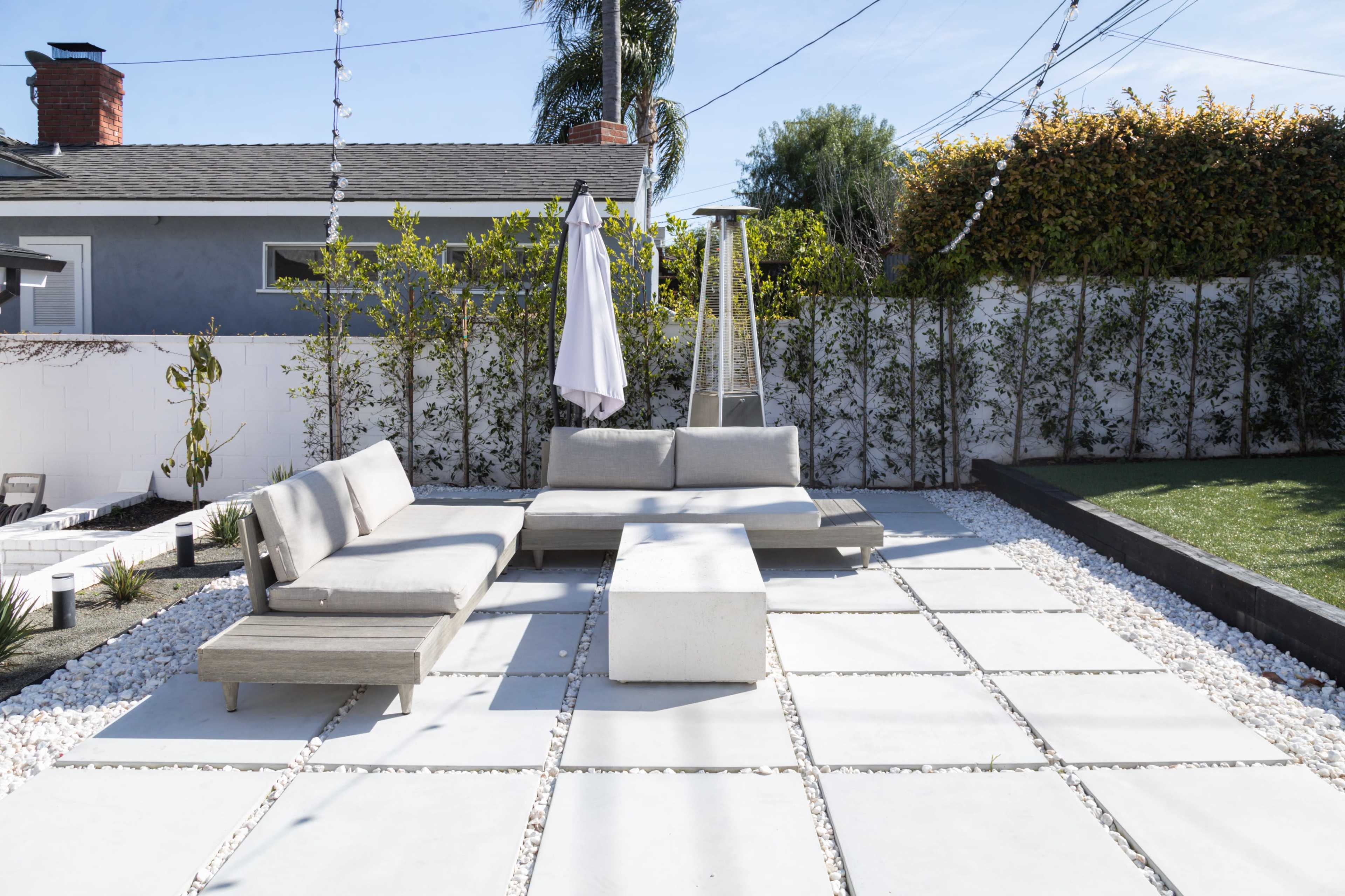 A modern outdoor patio with concrete slabs, a gray sectional sofa, a matching coffee table, and a large umbrella, surrounded by greenery and white fencing.