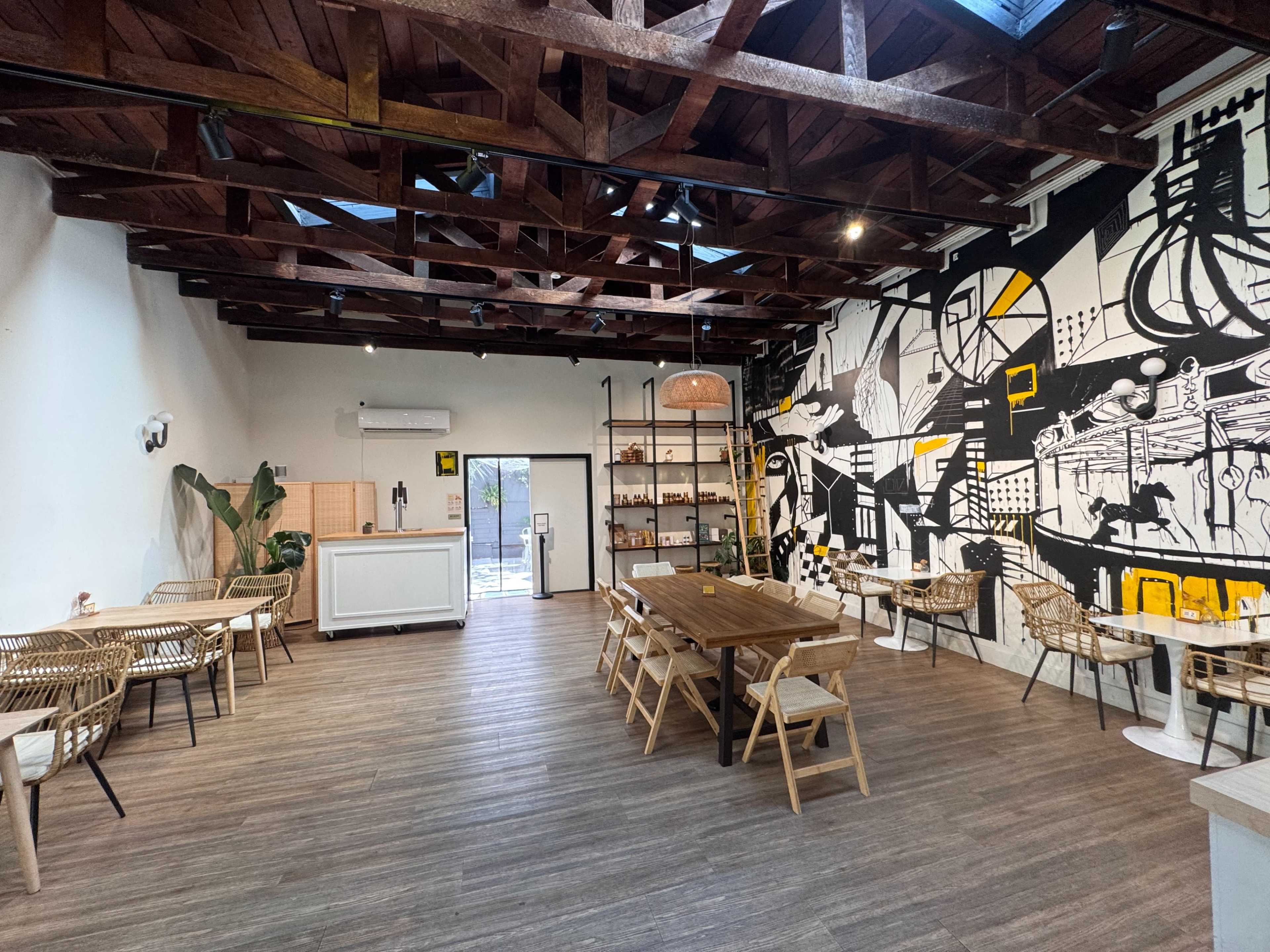 The image shows a modern café interior with wooden beams, a large mural on one wall, and various seating arrangements including tables and chairs.