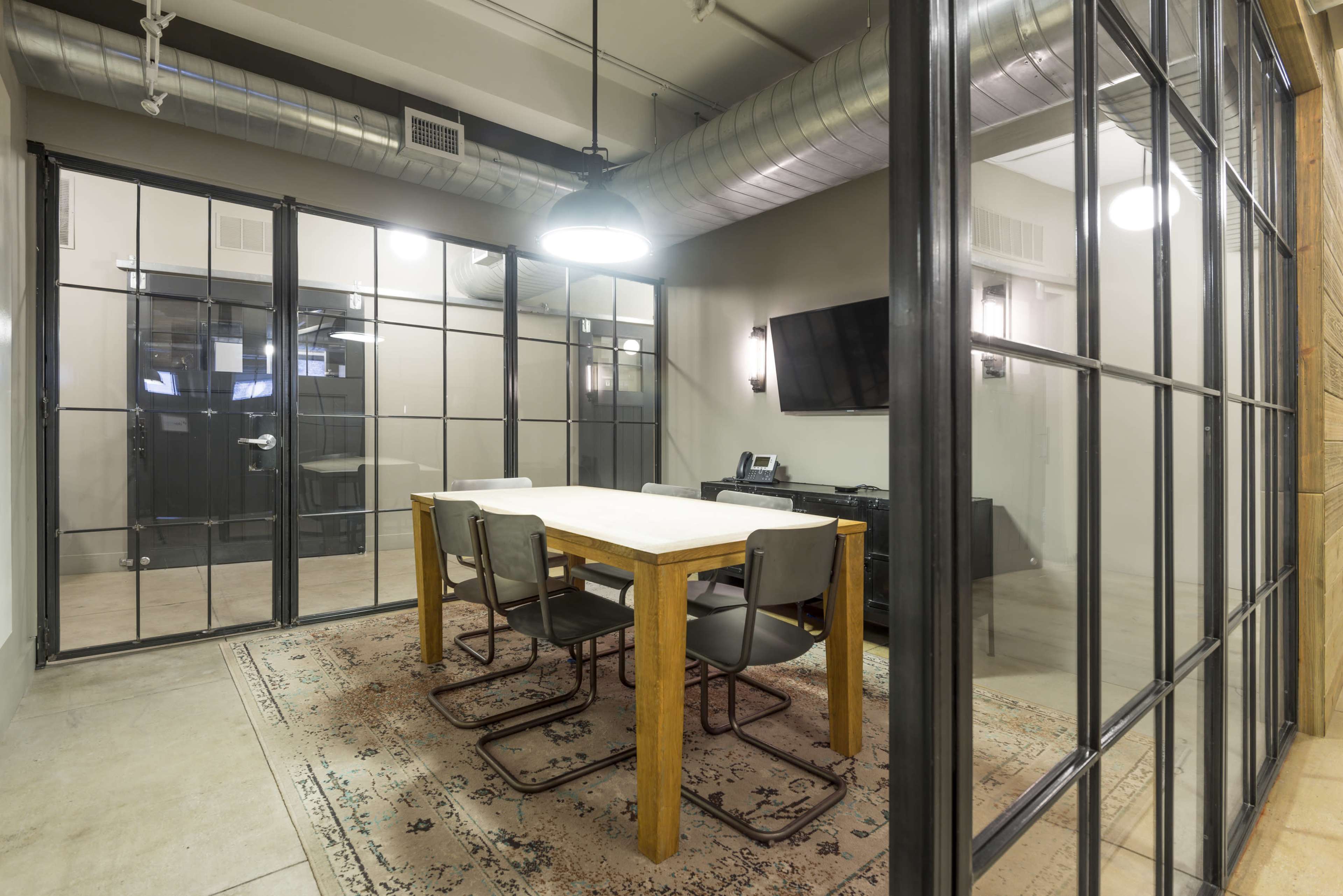 The image shows a modern conference room with a wooden table, four black chairs, and glass walls, featuring industrial-style lighting and a television on one wall.