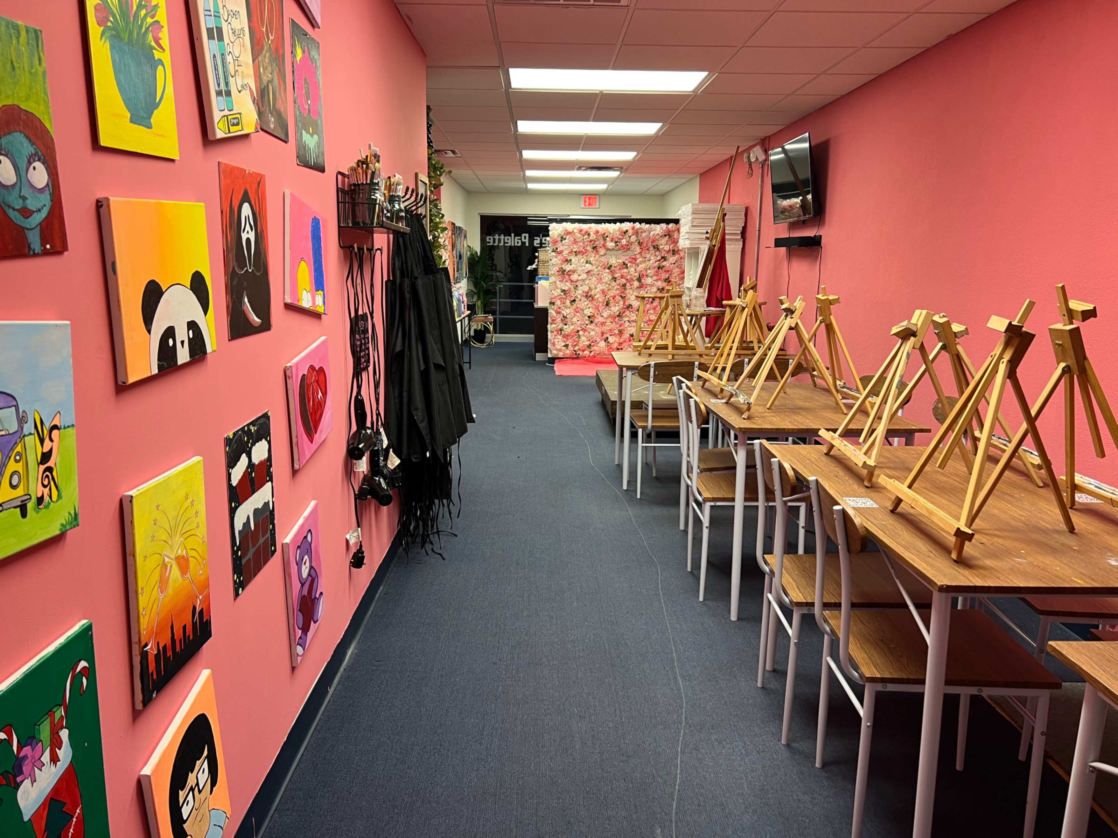 An art studio with pink walls, decorated with various paintings, featuring empty tables and easels arranged along the sides.