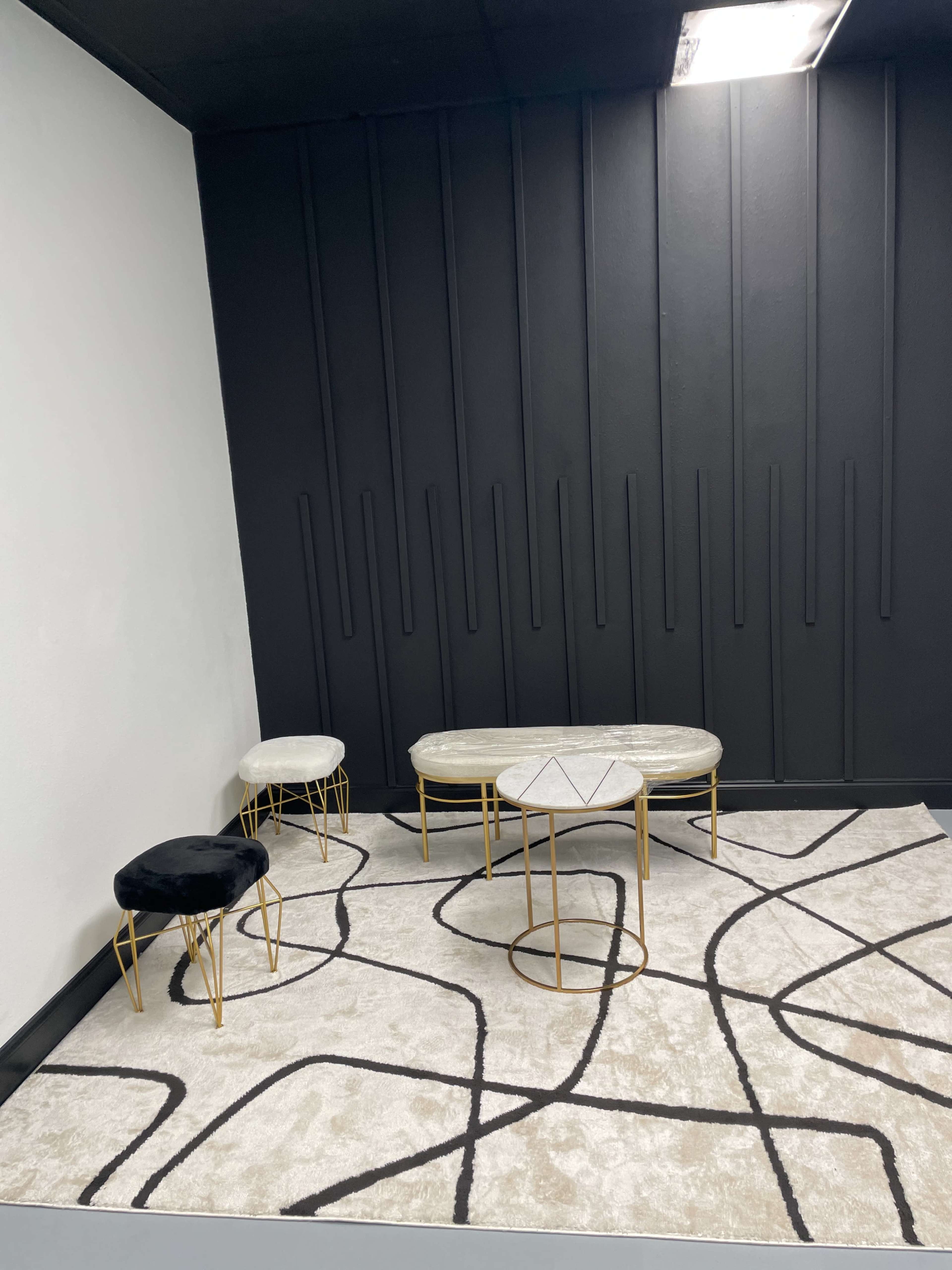 A minimalist interior space featuring a geometric patterned rug with a light color, a black wall, and several marble-topped tables with gold bases.