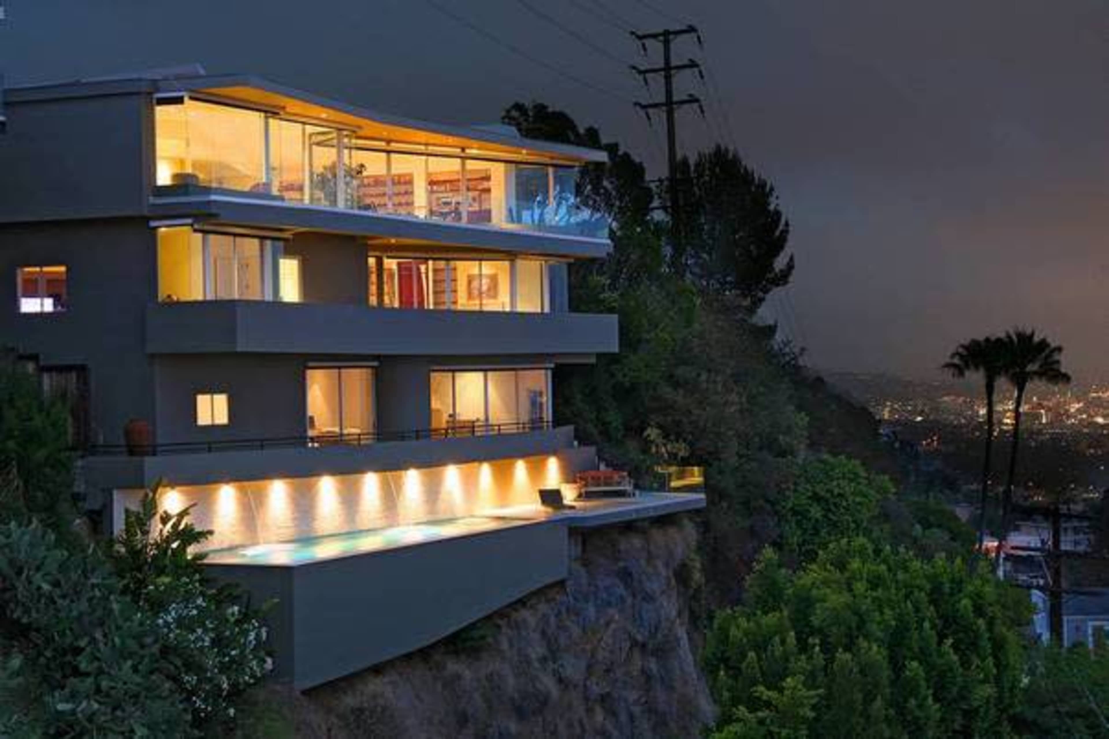 A modern three-level house with large windows overlooks a cityscape at night, featuring an illuminated infinity pool and greenery surrounding the property.