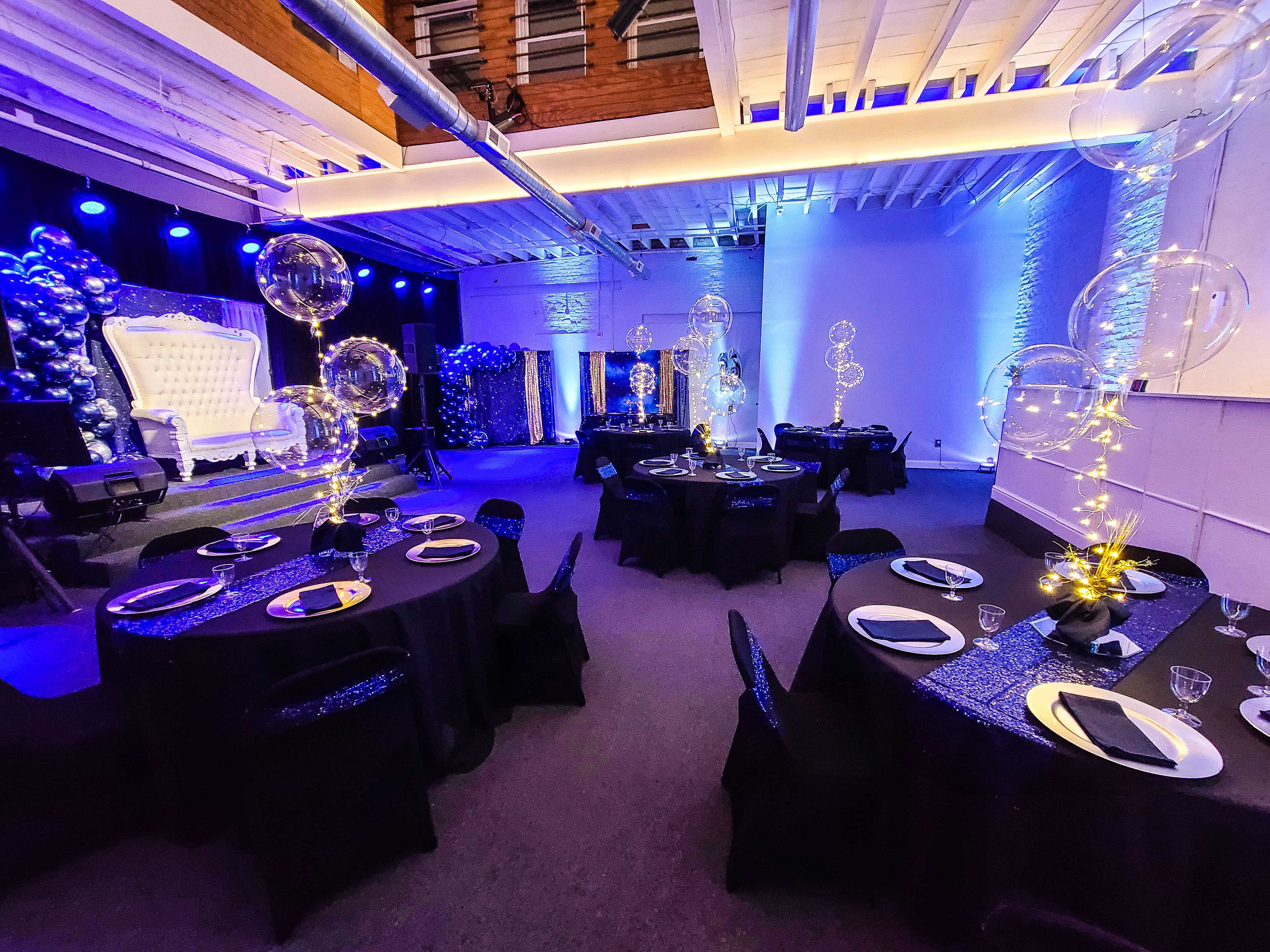 The venue features round tables with black tablecloths and elegant centerpieces, surrounded by balloons and decorative lights in a dimly lit setting.