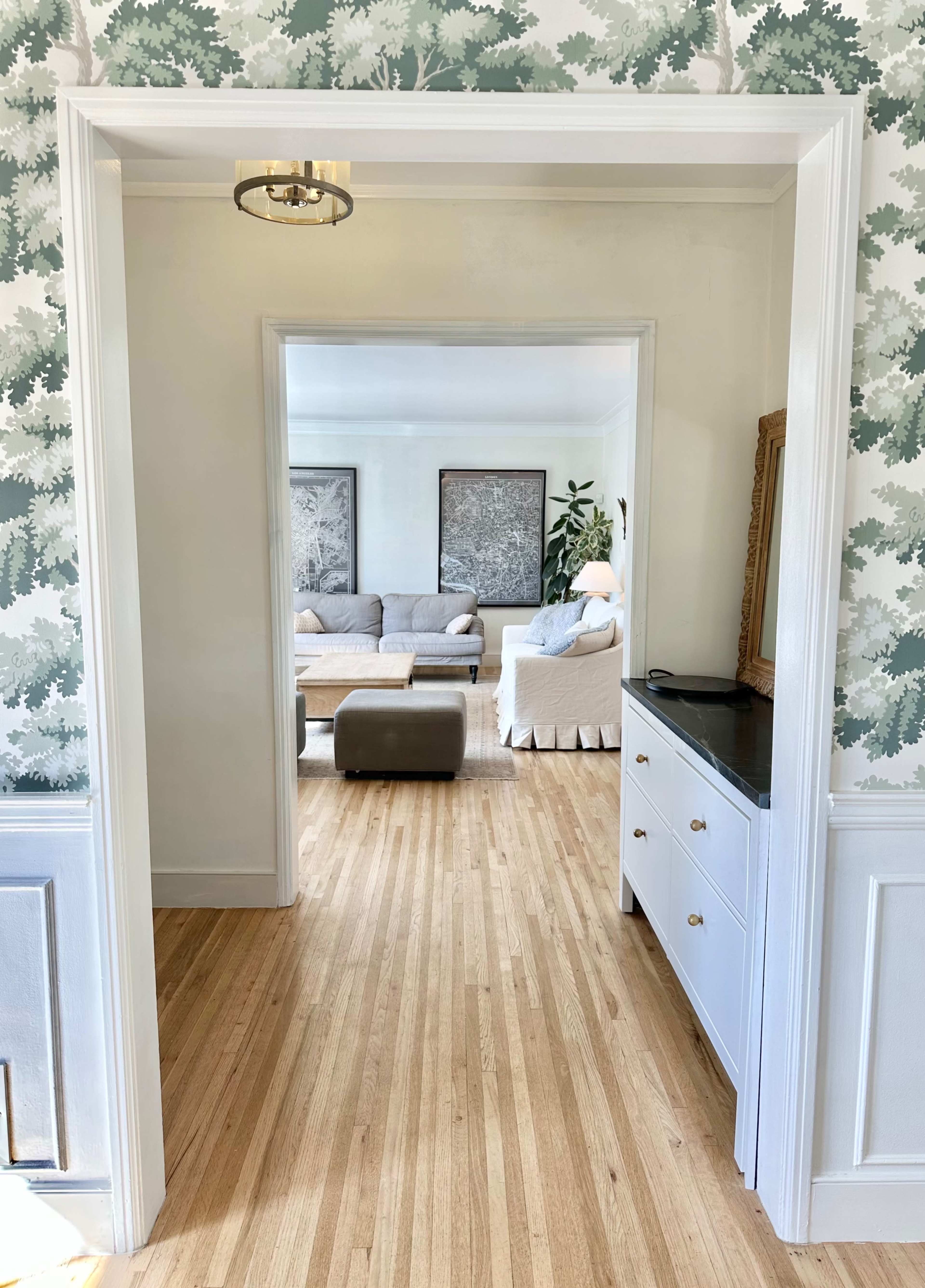 A doorway leads from a patterned wall into a spacious living room with a gray sofa, light-colored decor, and wooden flooring.