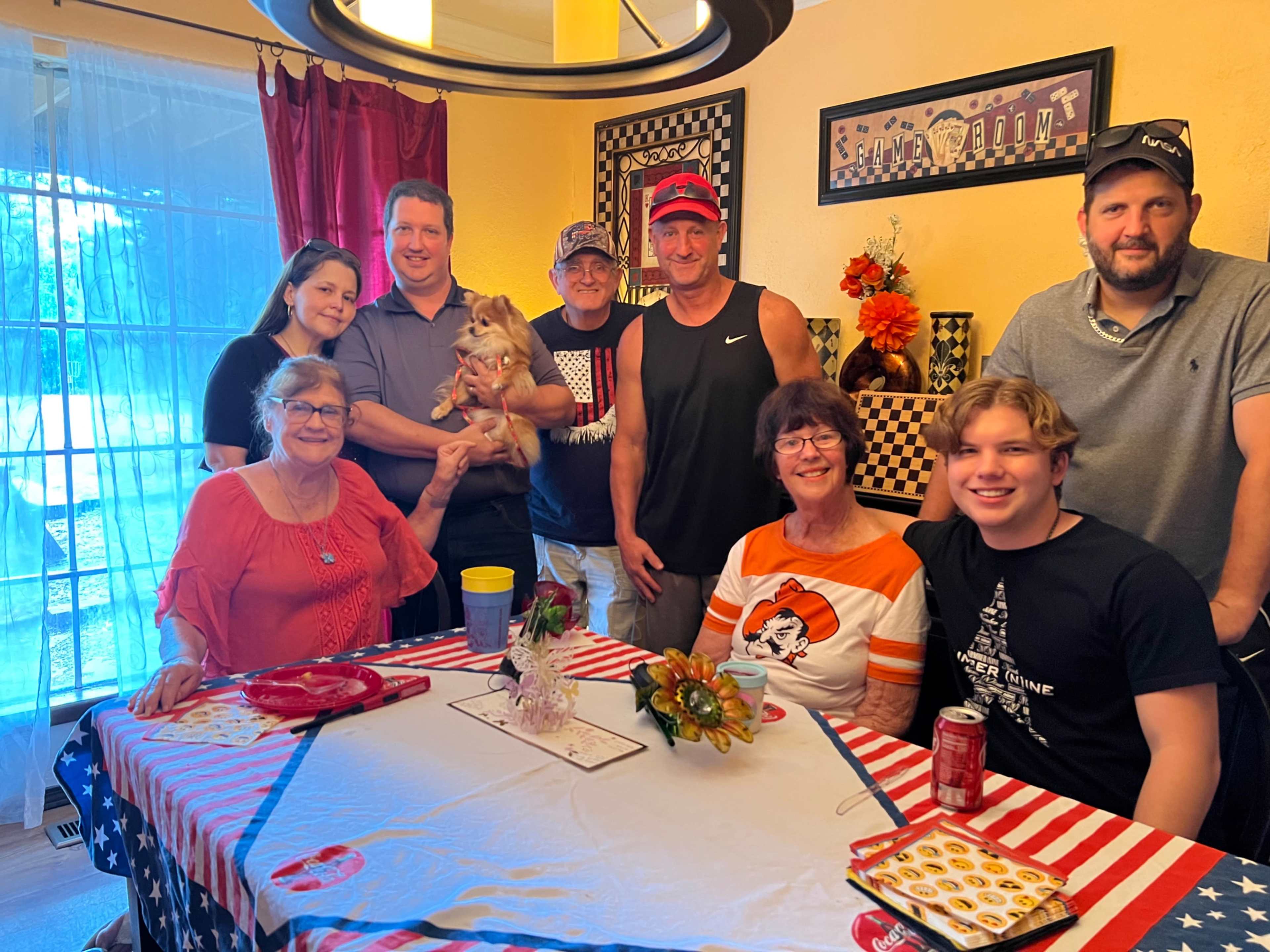 A group of eight people poses together in a dining room decorated with checkered patterns and American flags, with a small dog held by one person.