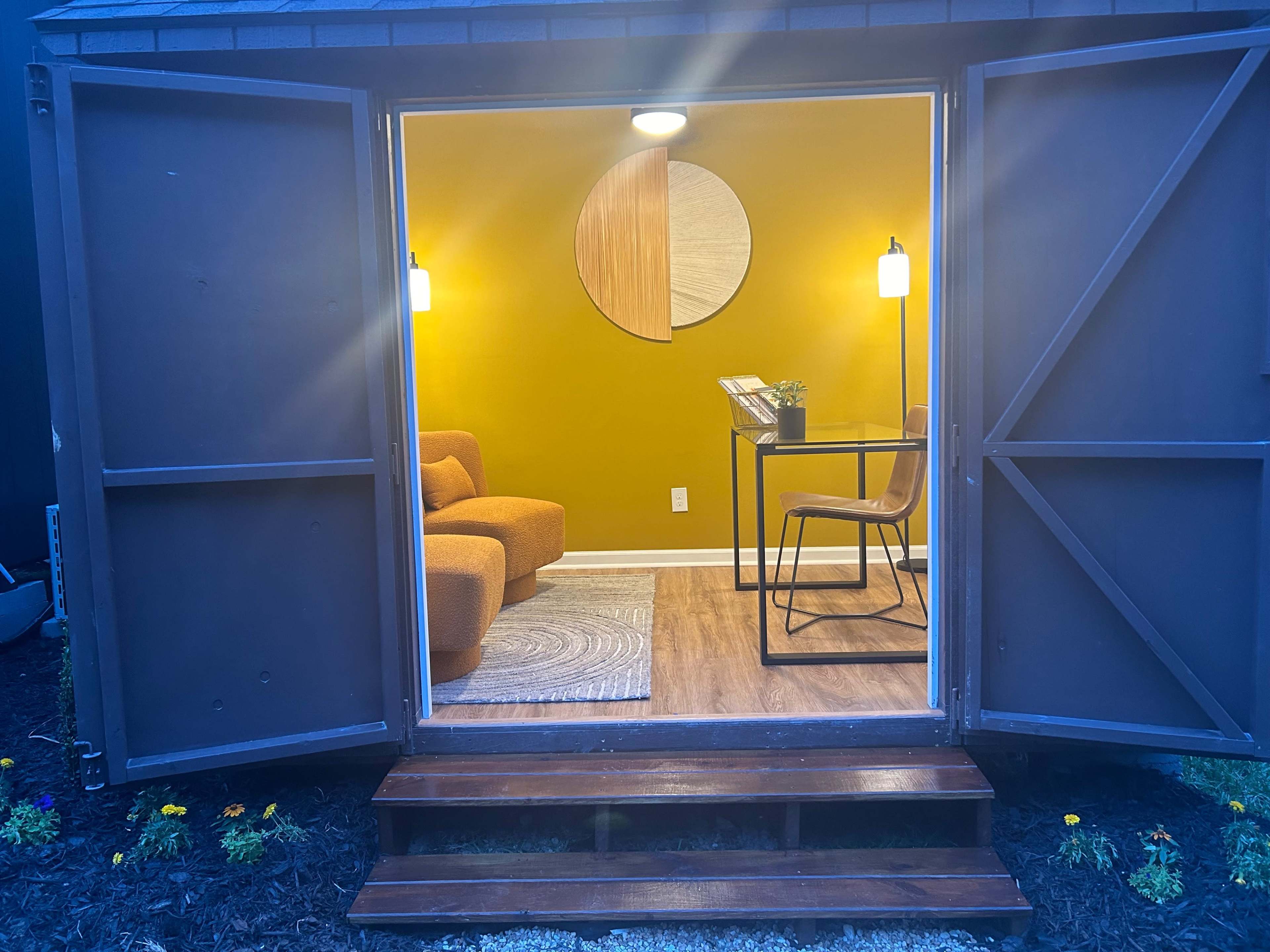 An open storage shed revealing a cozy interior with a yellow wall, a round decorative piece, a sofa, a desk, and two pendant lights.