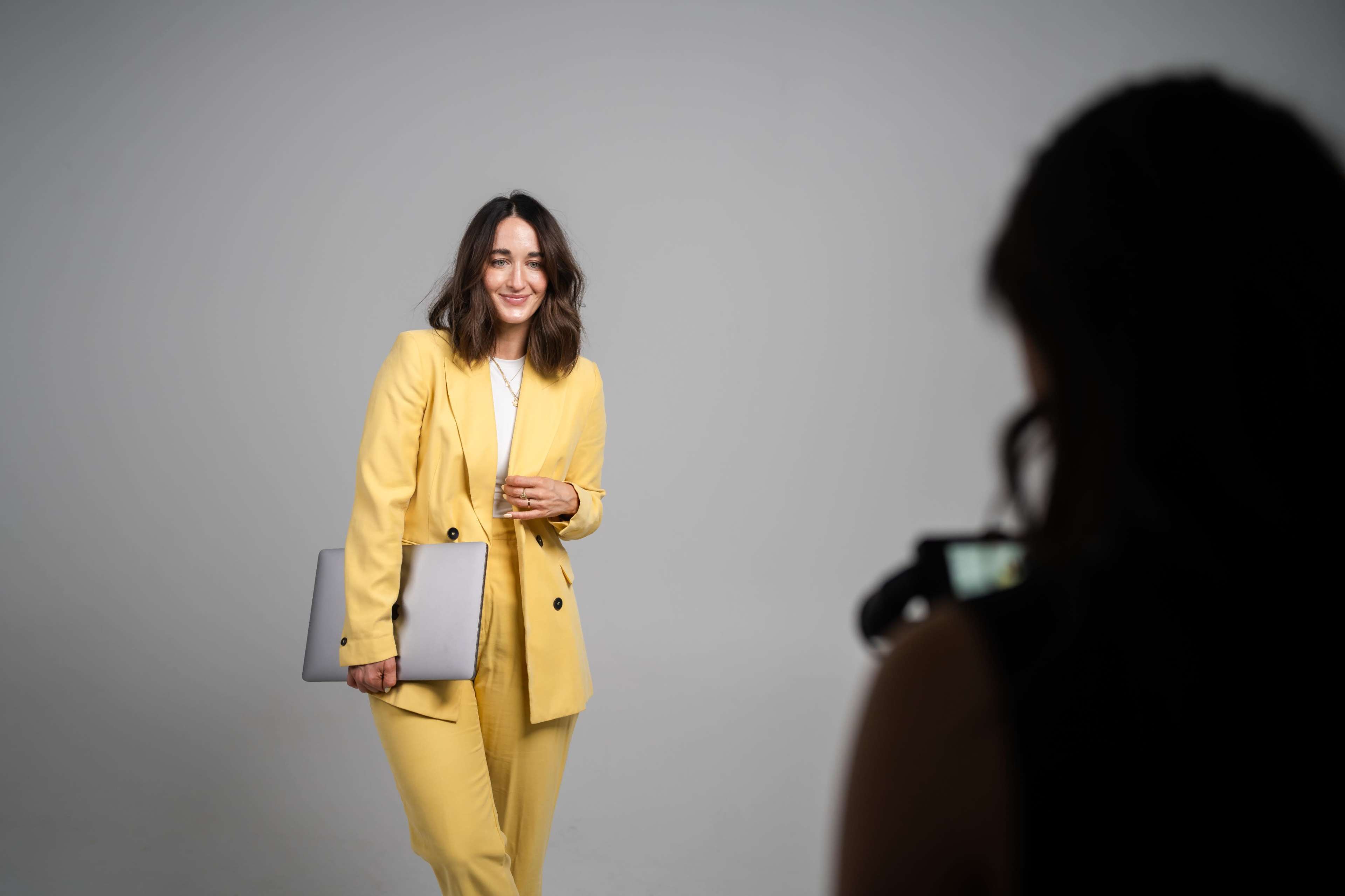 A woman in a yellow suit poses while holding a laptop and smiling at a photographer off-camera.