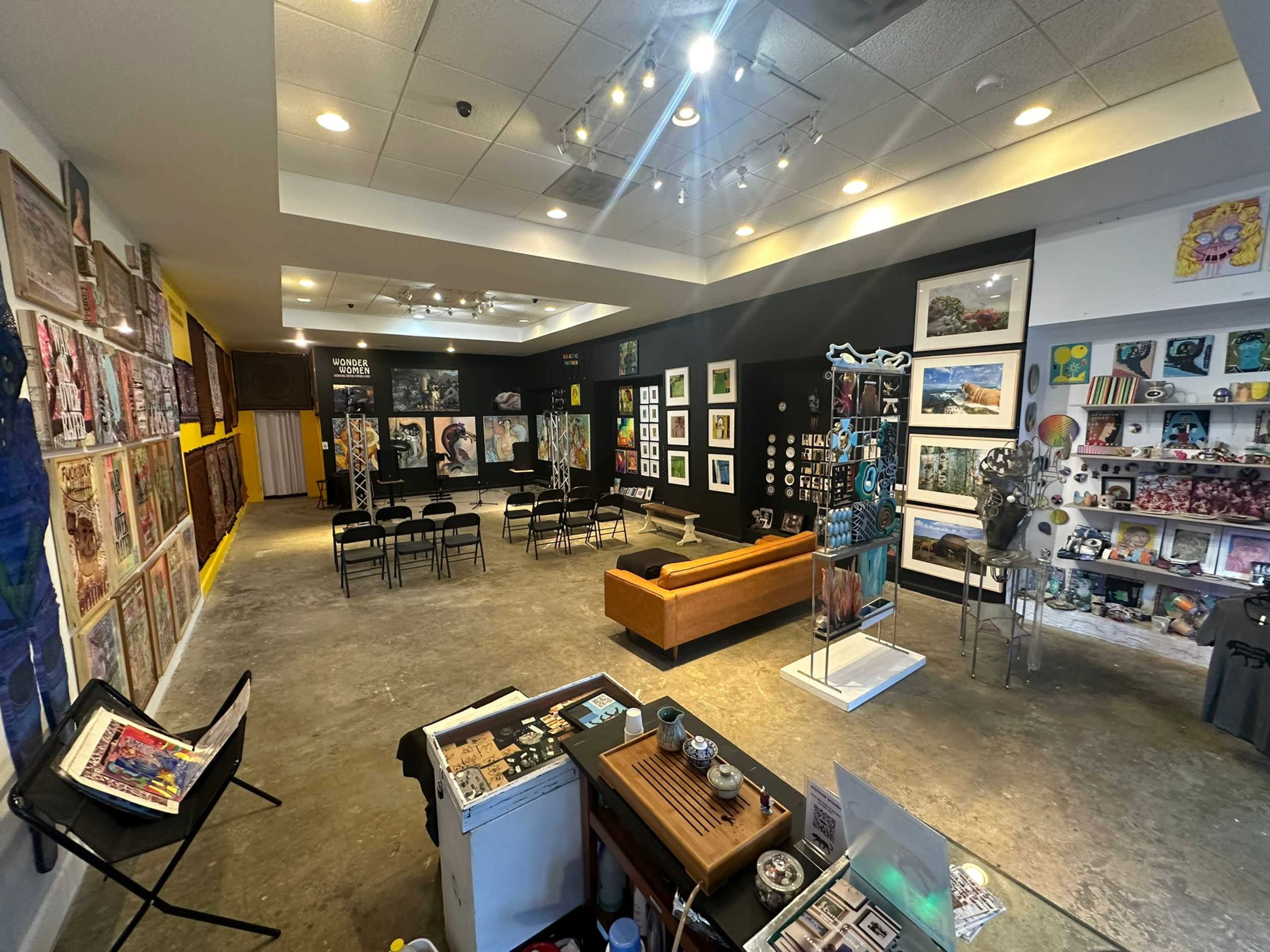 An art gallery featuring a variety of framed artworks on the walls, a seating area with black chairs, and a wooden couch in a well-lit, spacious room.