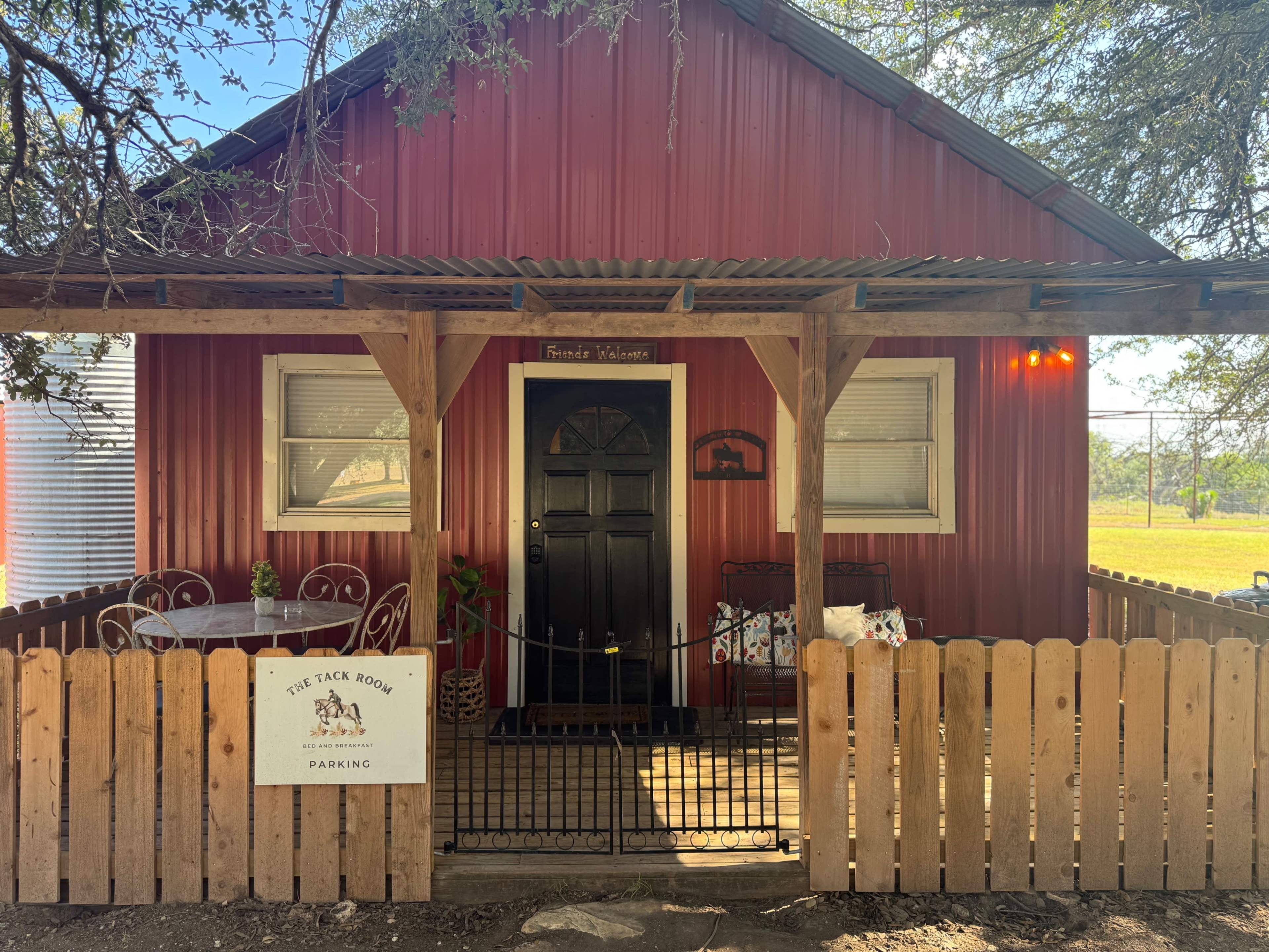 The image shows a small red cabin with a black door and a covered porch, surrounded by a wooden fence and featuring a sign that reads "The Tack Room Parking."