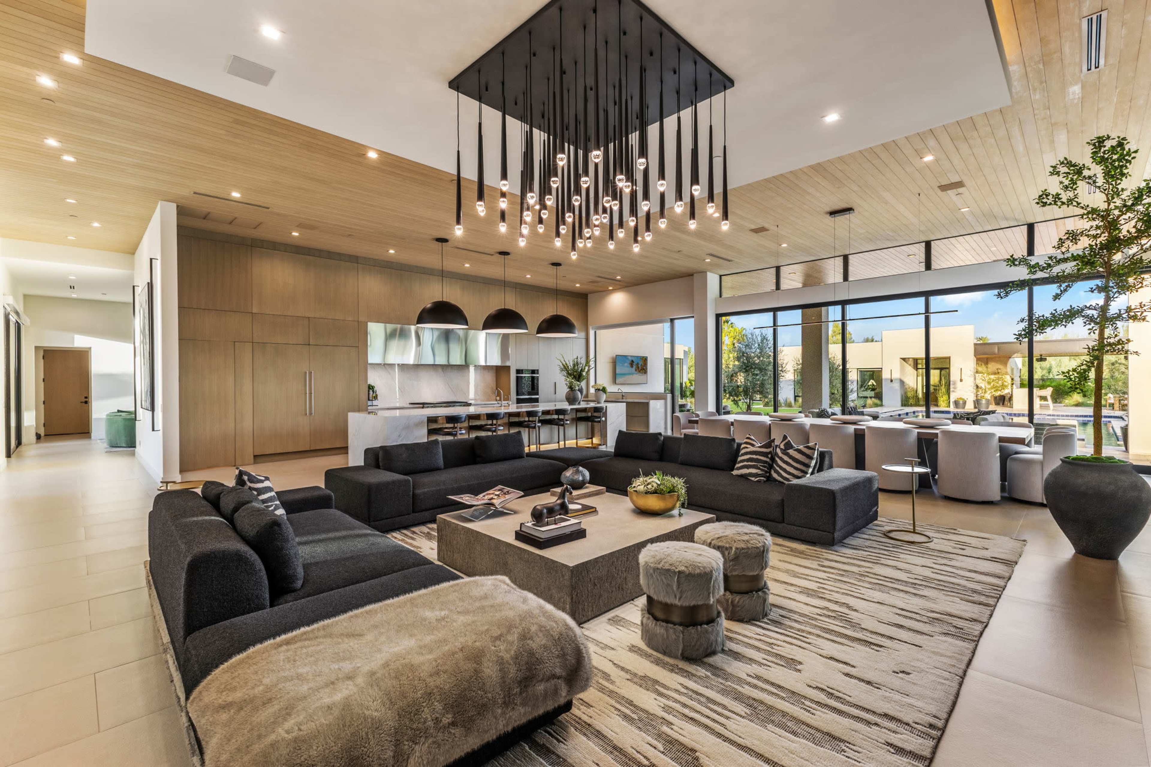 The spacious living room features a large sectional sofa, a decorative coffee table, and a modern chandelier, with large windows providing natural light and views of the outdoors.