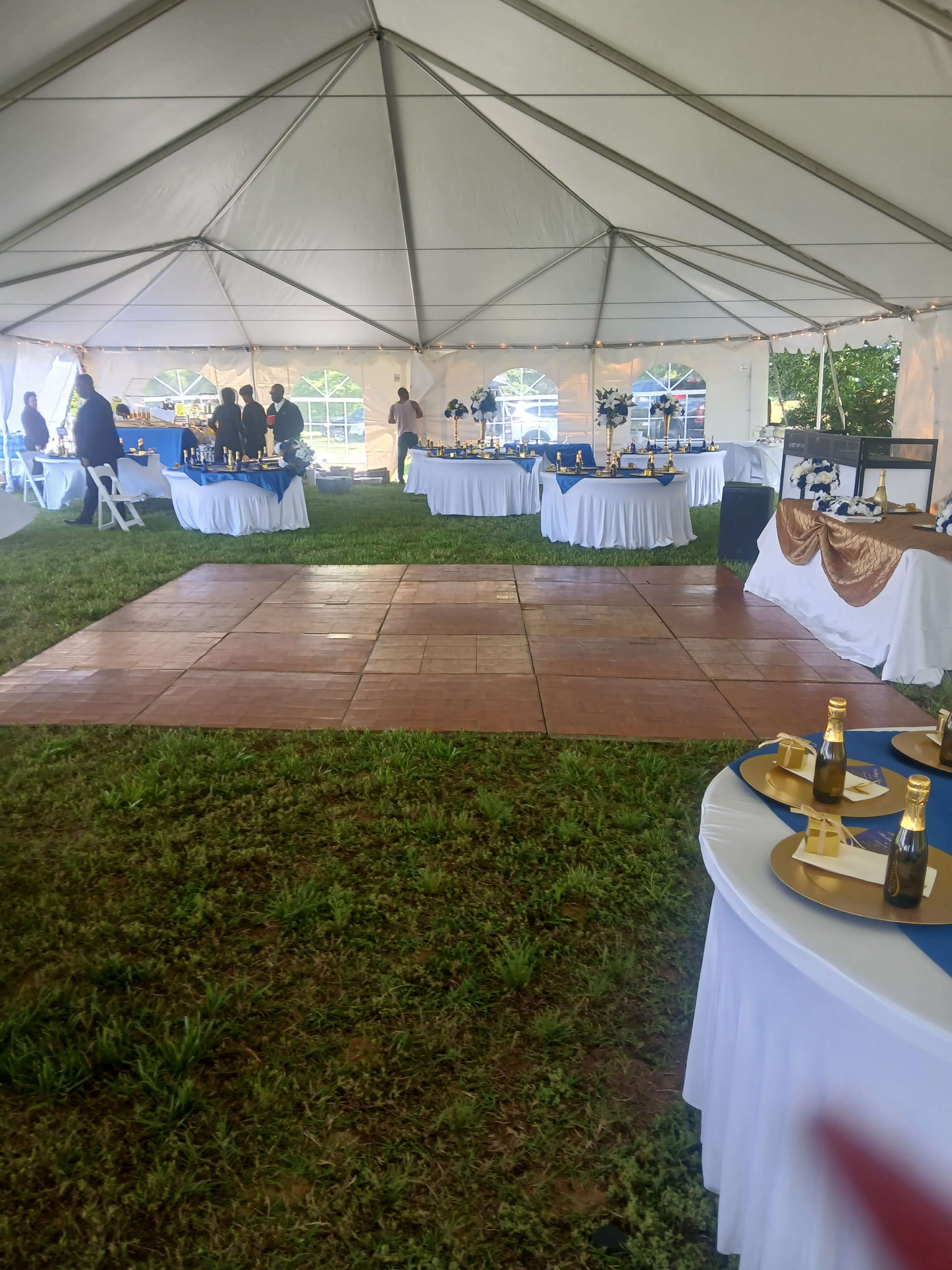 A large white tent is set up on grass with several tables covered in white tablecloths, some adorned with decorations, and a small dance floor in the center.