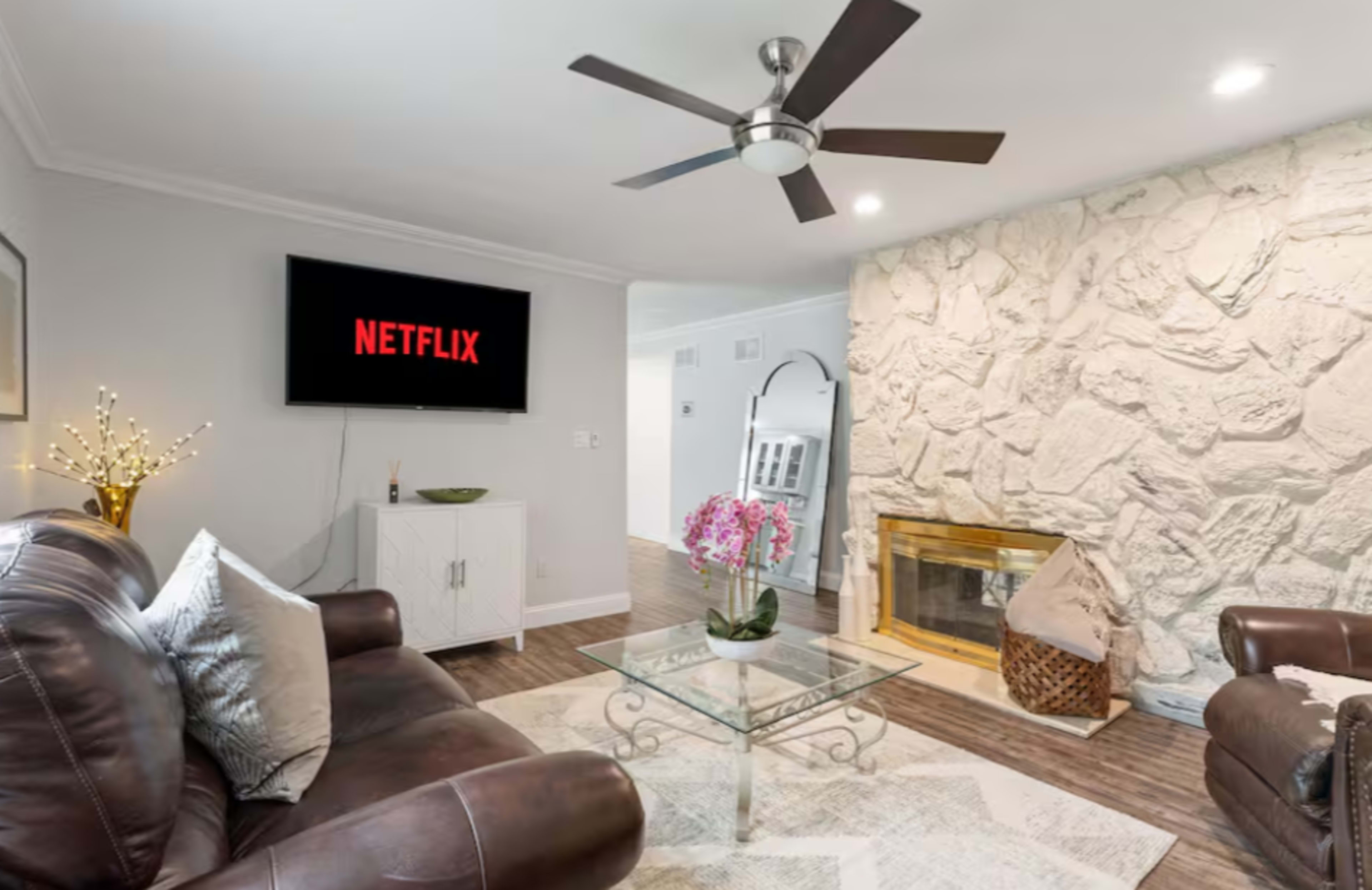 The living room features a leather sofa, a glass coffee table, a wall-mounted TV displaying the Netflix logo, and a stone fireplace.