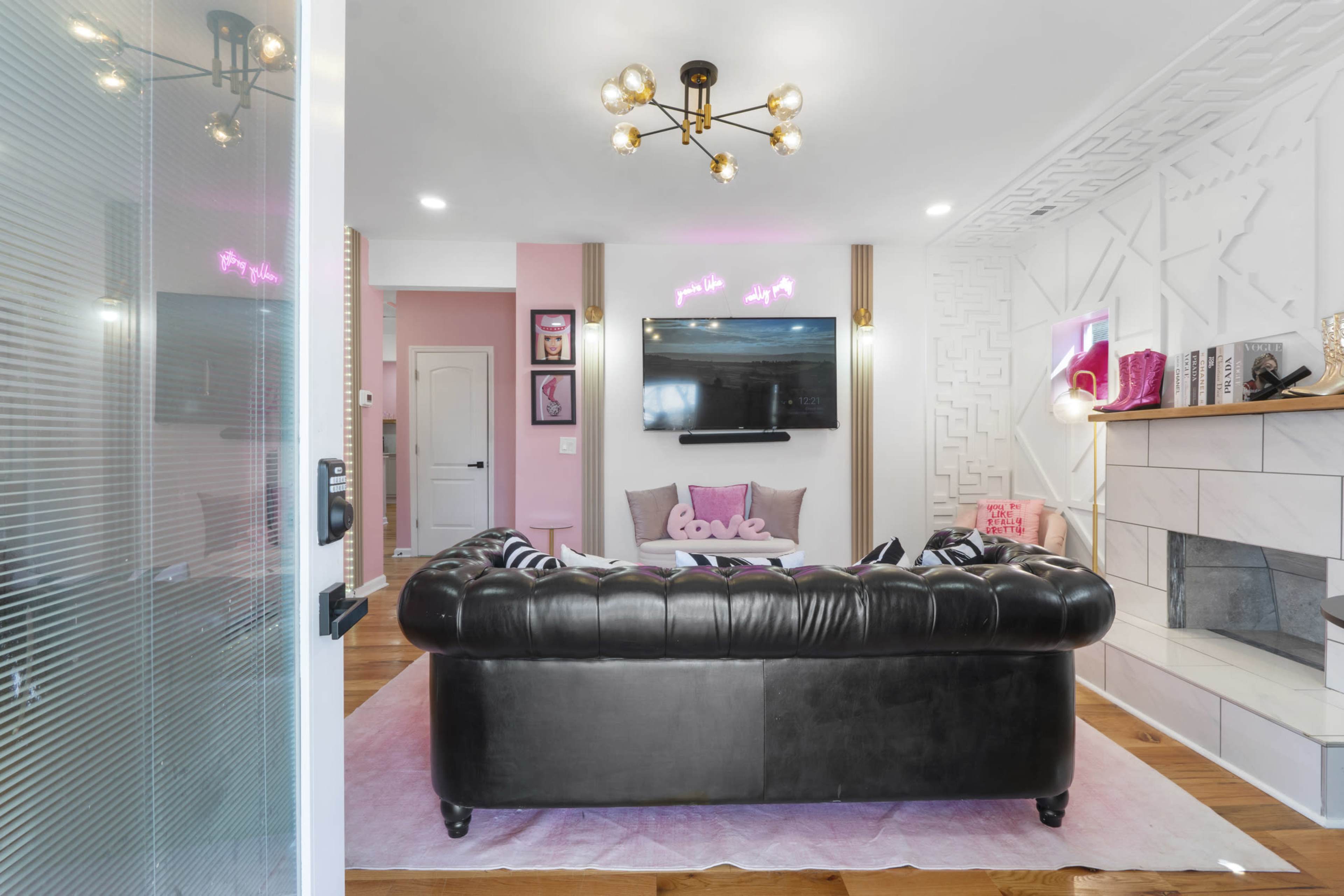 The image shows a modern living room featuring a black leather chesterfield sofa, a wall-mounted television, and various decorative elements in shades of pink.