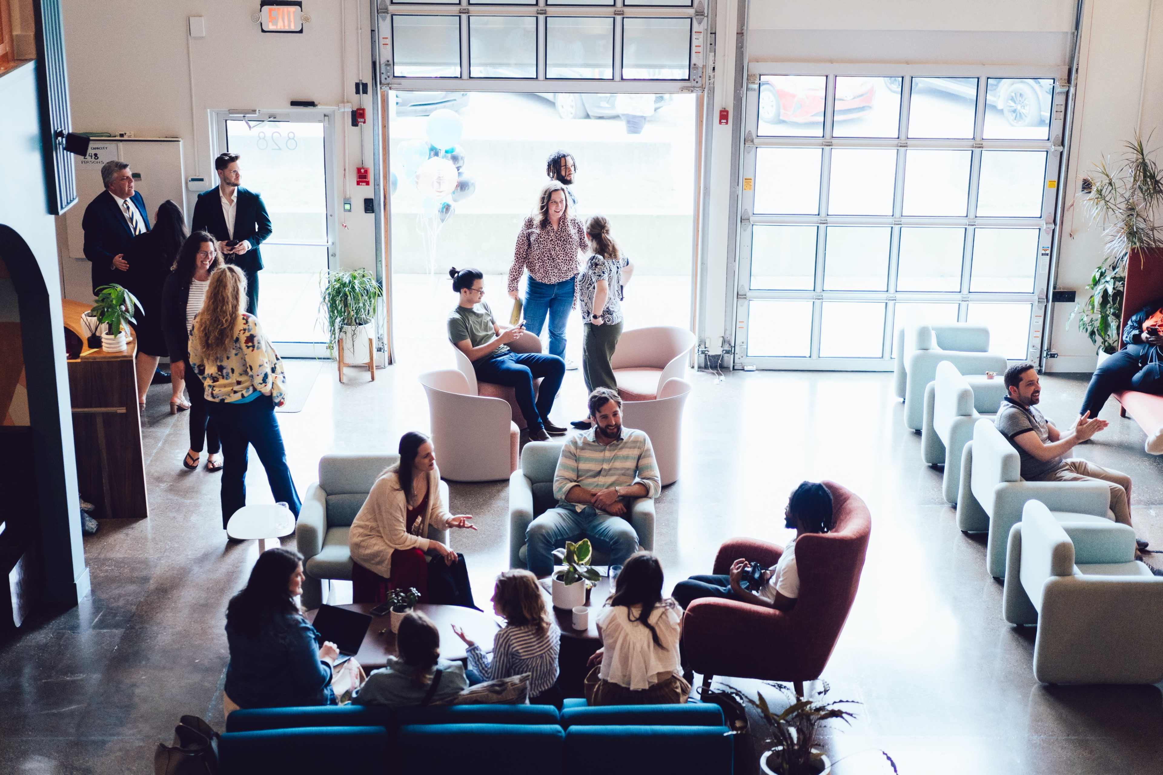 A group of people socializes in a modern open space with large windows and various seating arrangements.