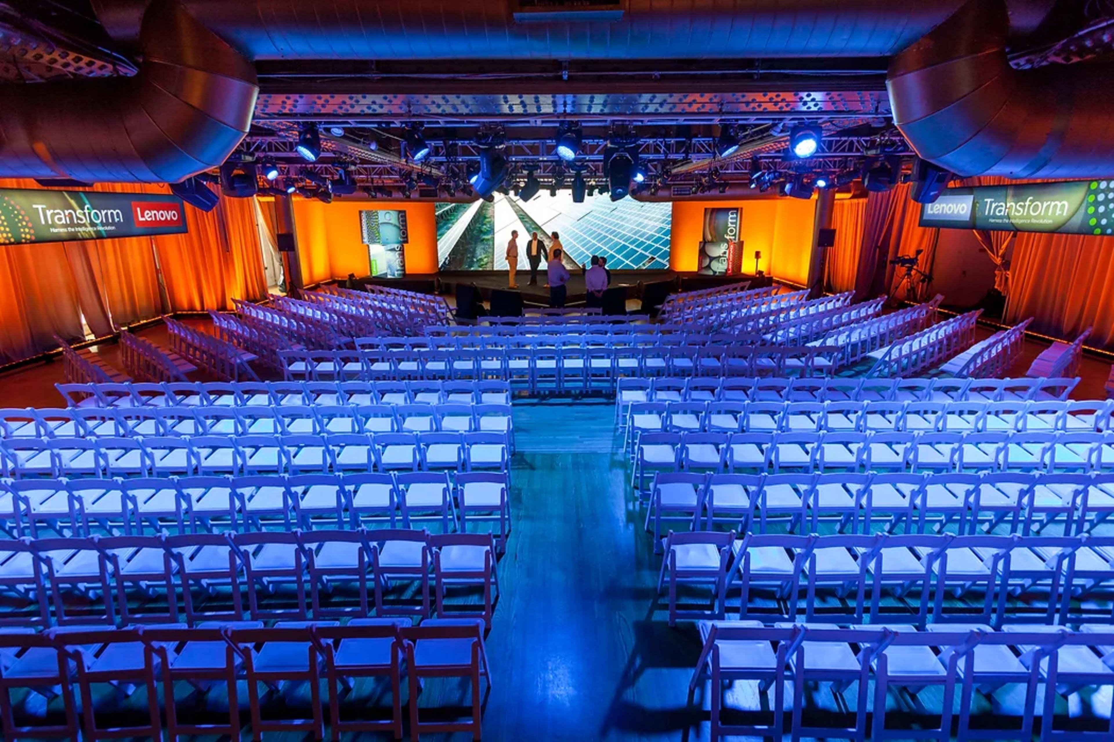 A spacious indoor venue set up with rows of white chairs facing a stage adorned with screens and illuminated by orange and blue lighting.