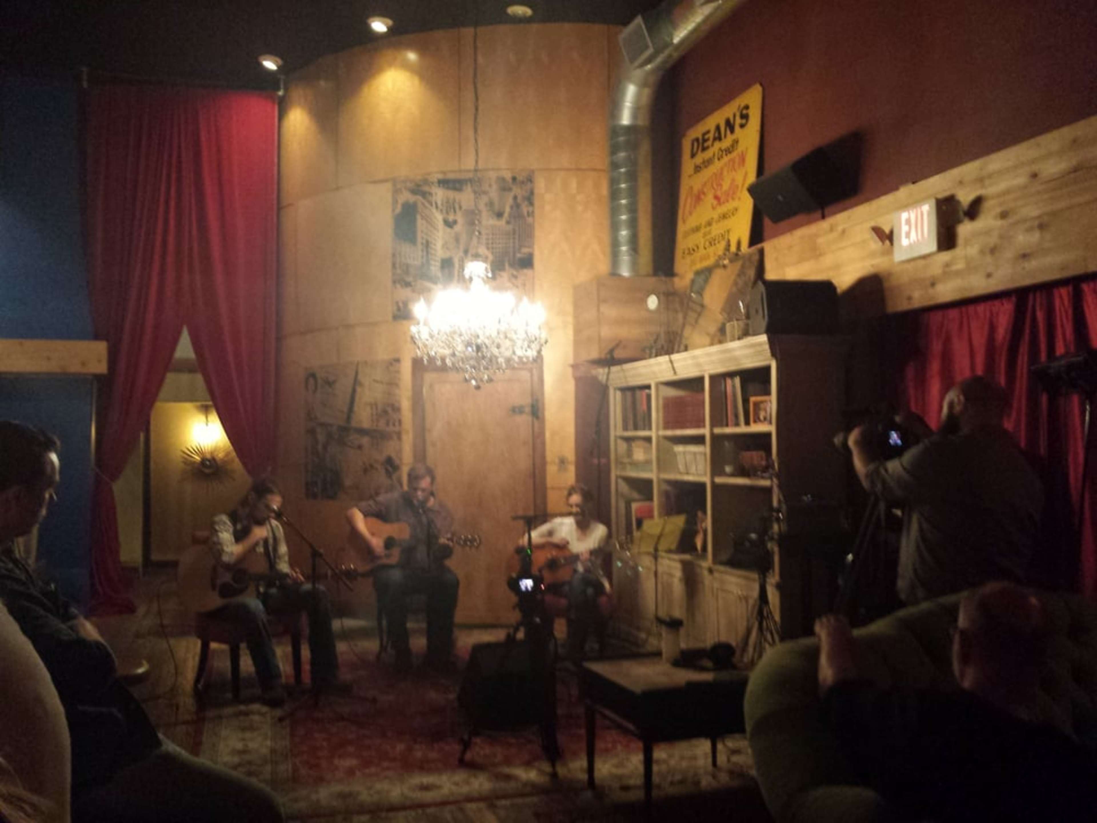 A small crowd watches three musicians perform in an intimate venue with a chandelier and vintage decor.