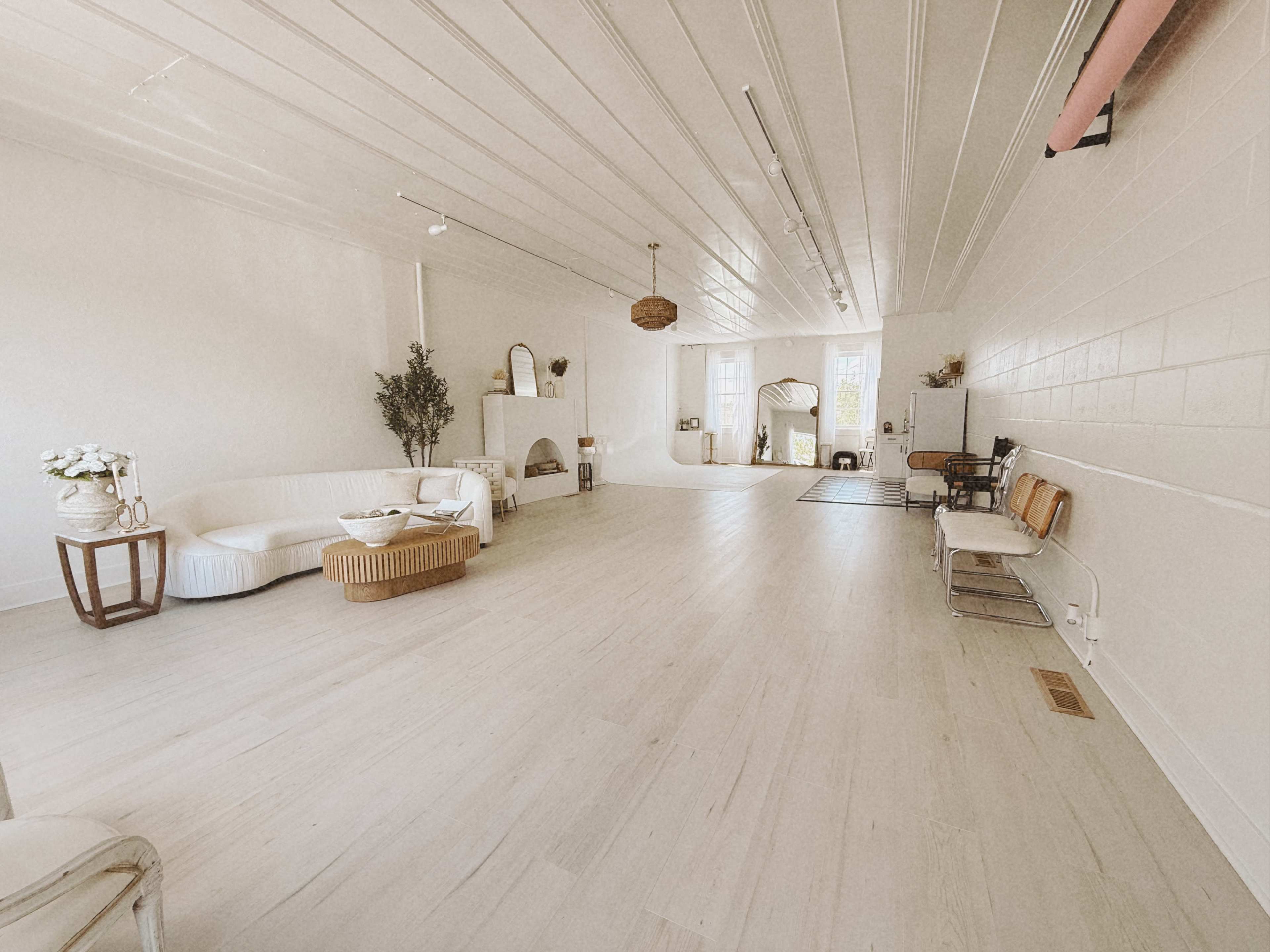 The image shows a spacious, minimally decorated interior with light wooden floors, white walls, and various seating arrangements.