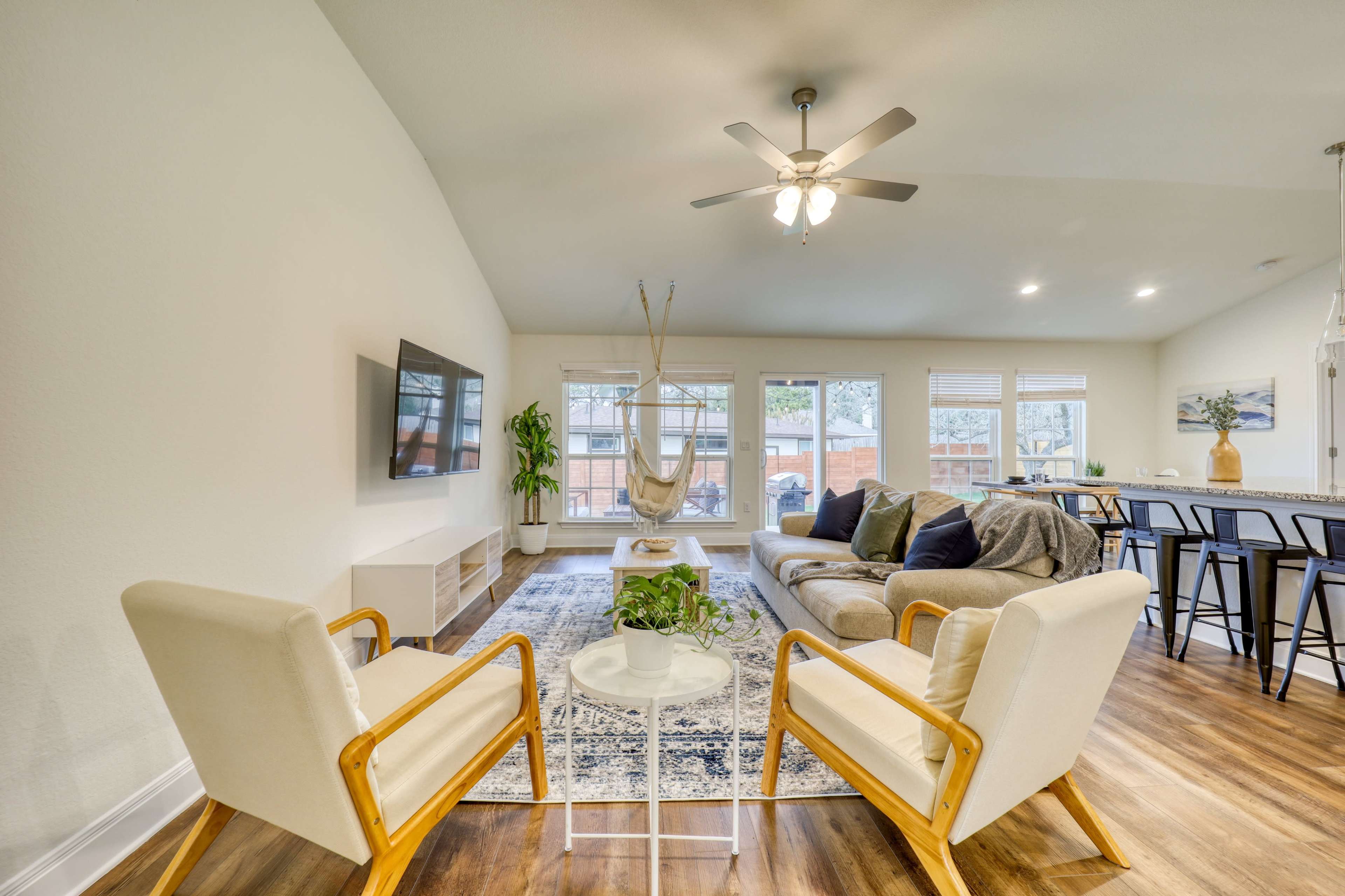 A modern living room with a gray sofa, two cream-colored armchairs, a hanging chair, a television mounted on the wall, and large windows that provide natural light.