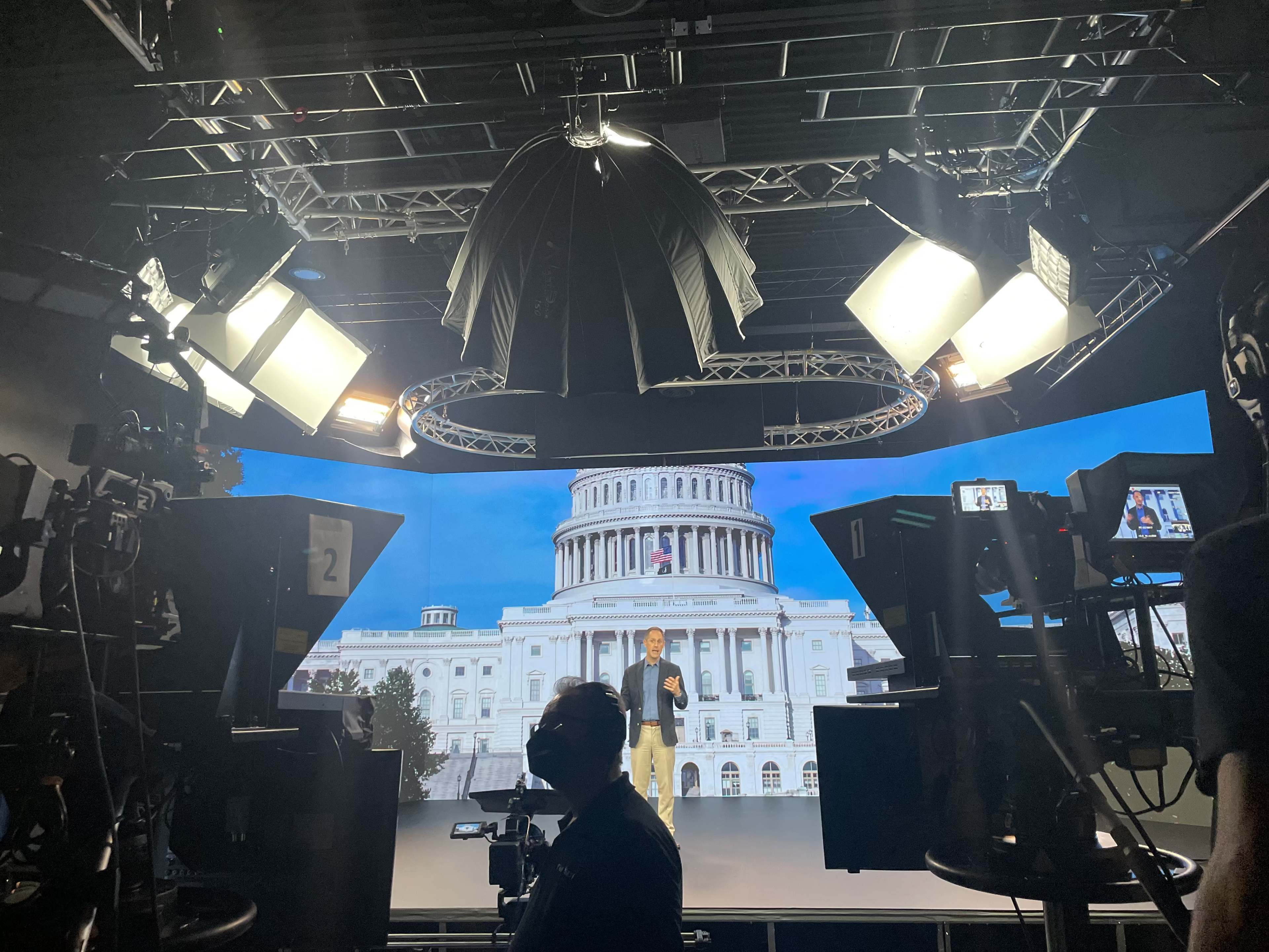 A man stands on a stage in a studio with the United States Capitol building projected in the background, surrounded by studio lights and cameras.