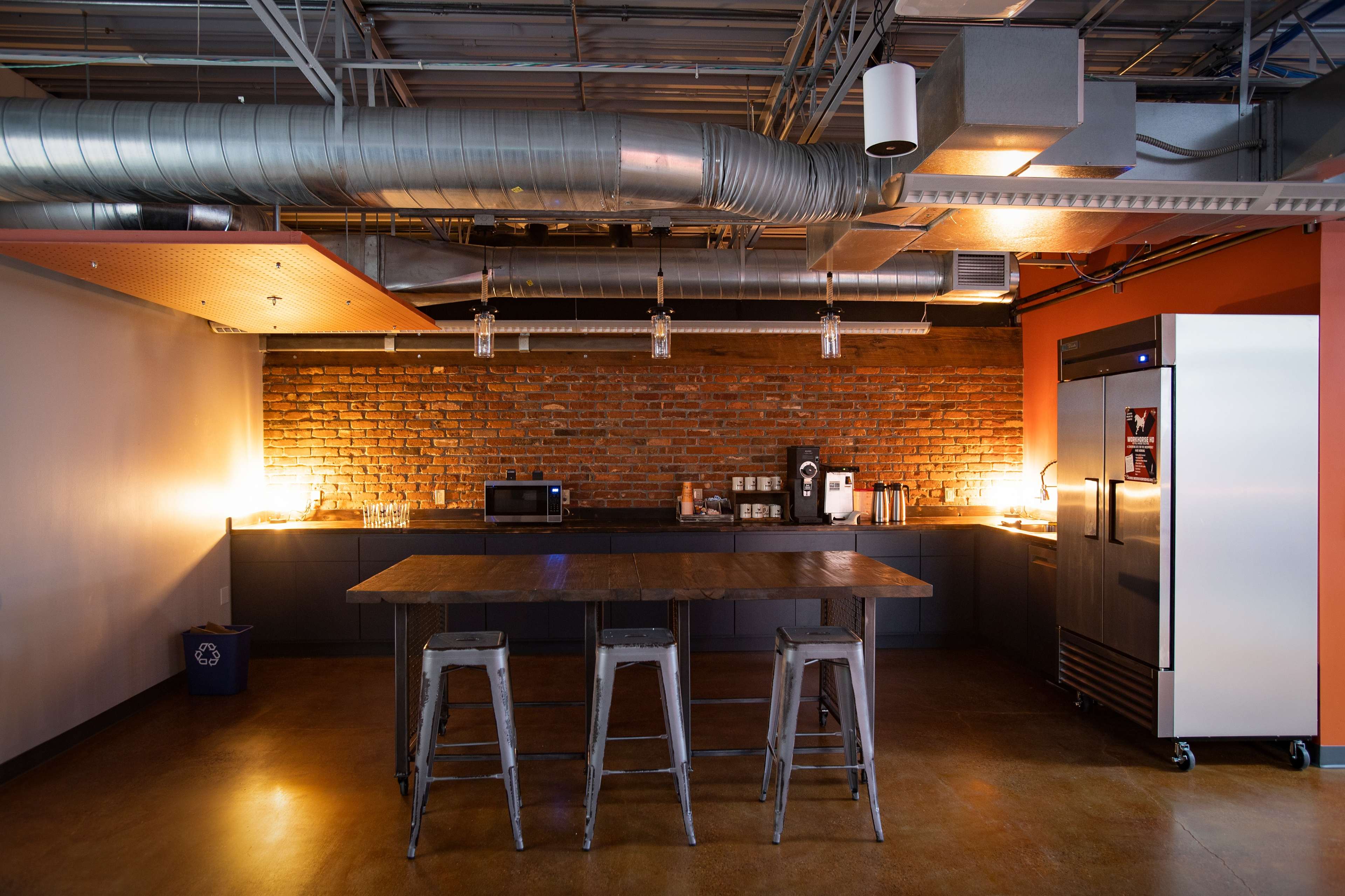 The image shows a modern kitchen area with a brick wall, stainless steel appliances, a large table, and metal stools.