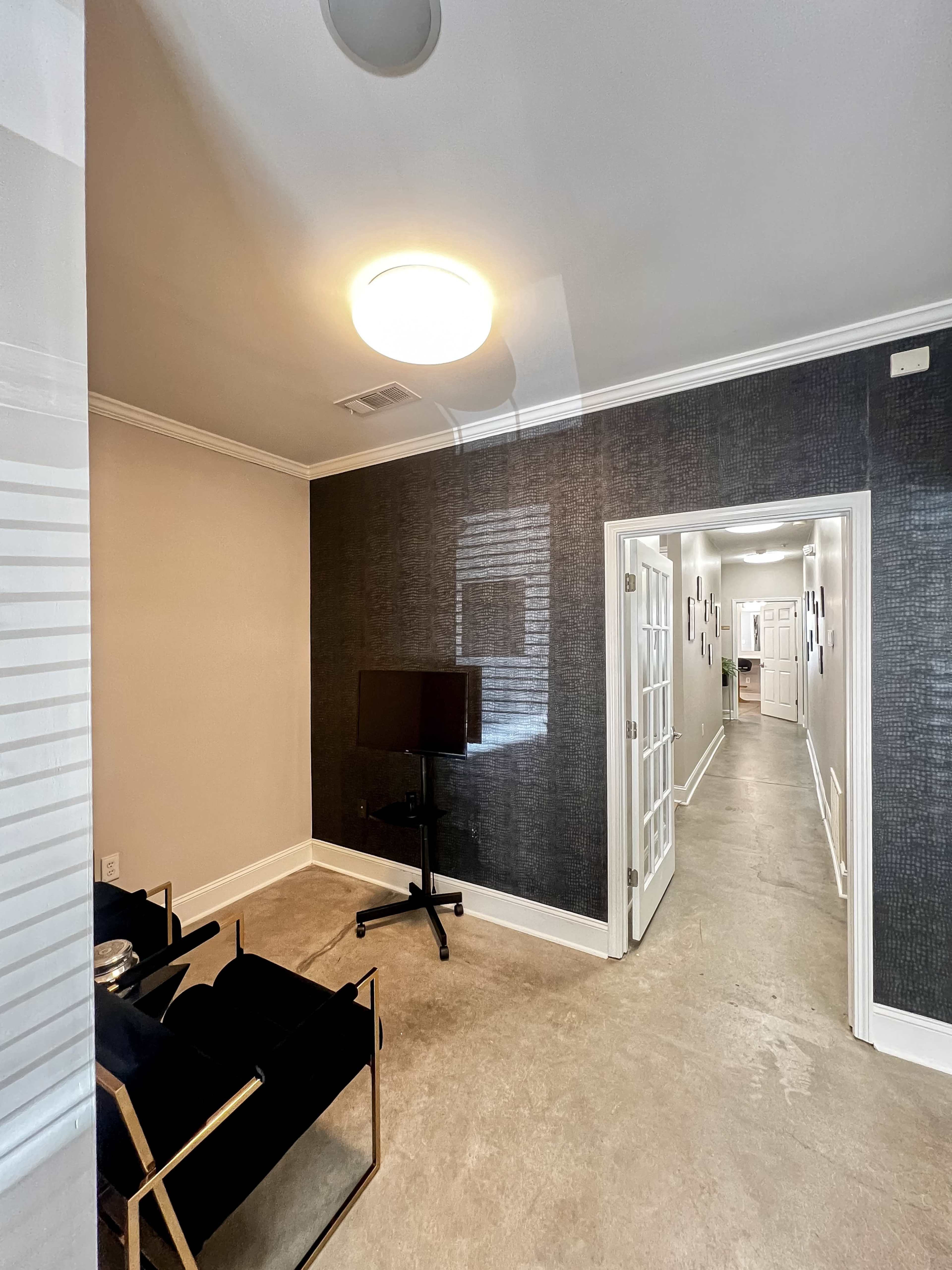 A minimalist interior space featuring a hallway with white doors on one side and a small sitting area with black chairs and a television on a stand.