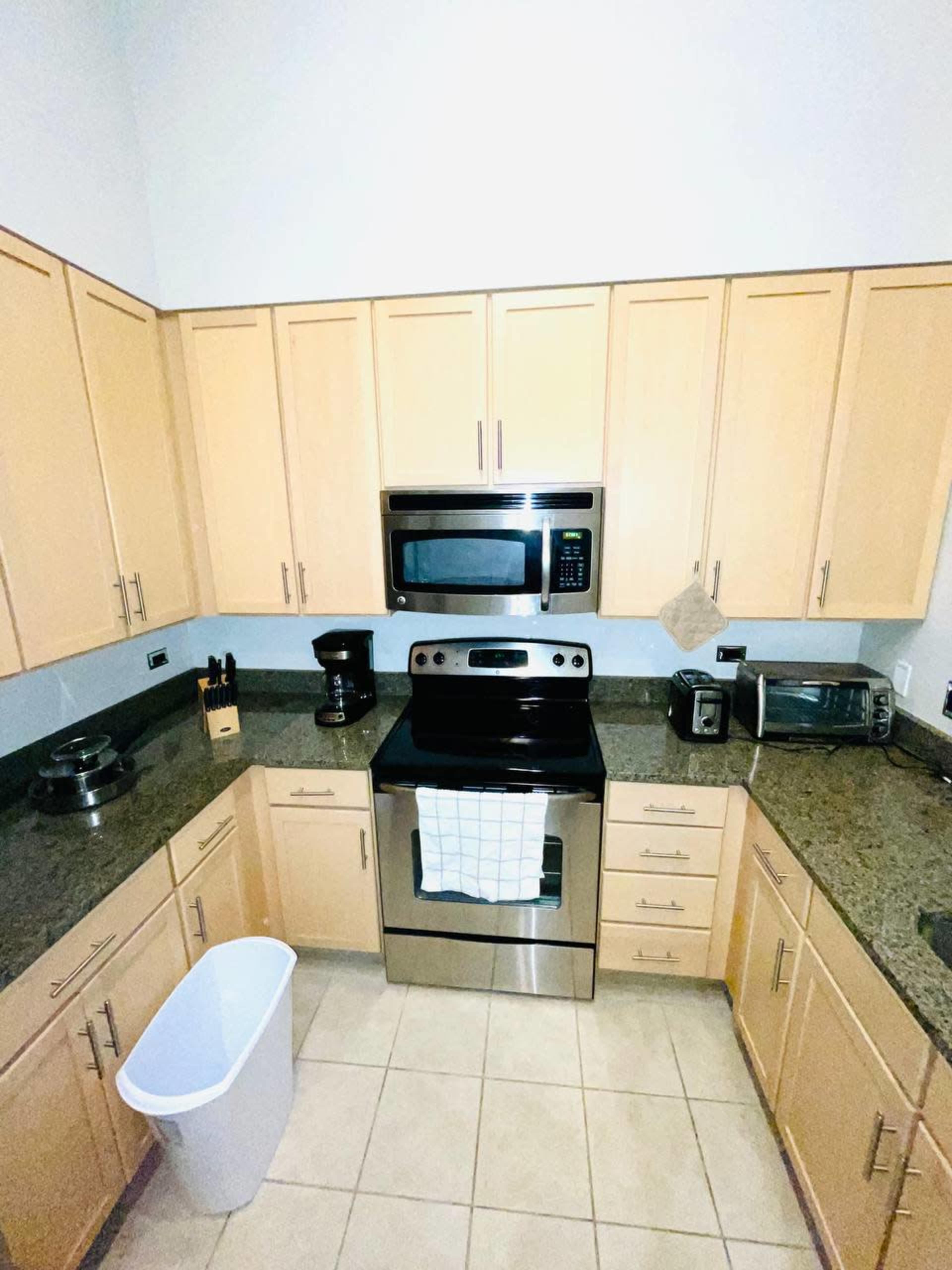 A kitchen with light wooden cabinetry, a stainless steel stove and microwave, and a white wastebasket on the tiled floor.