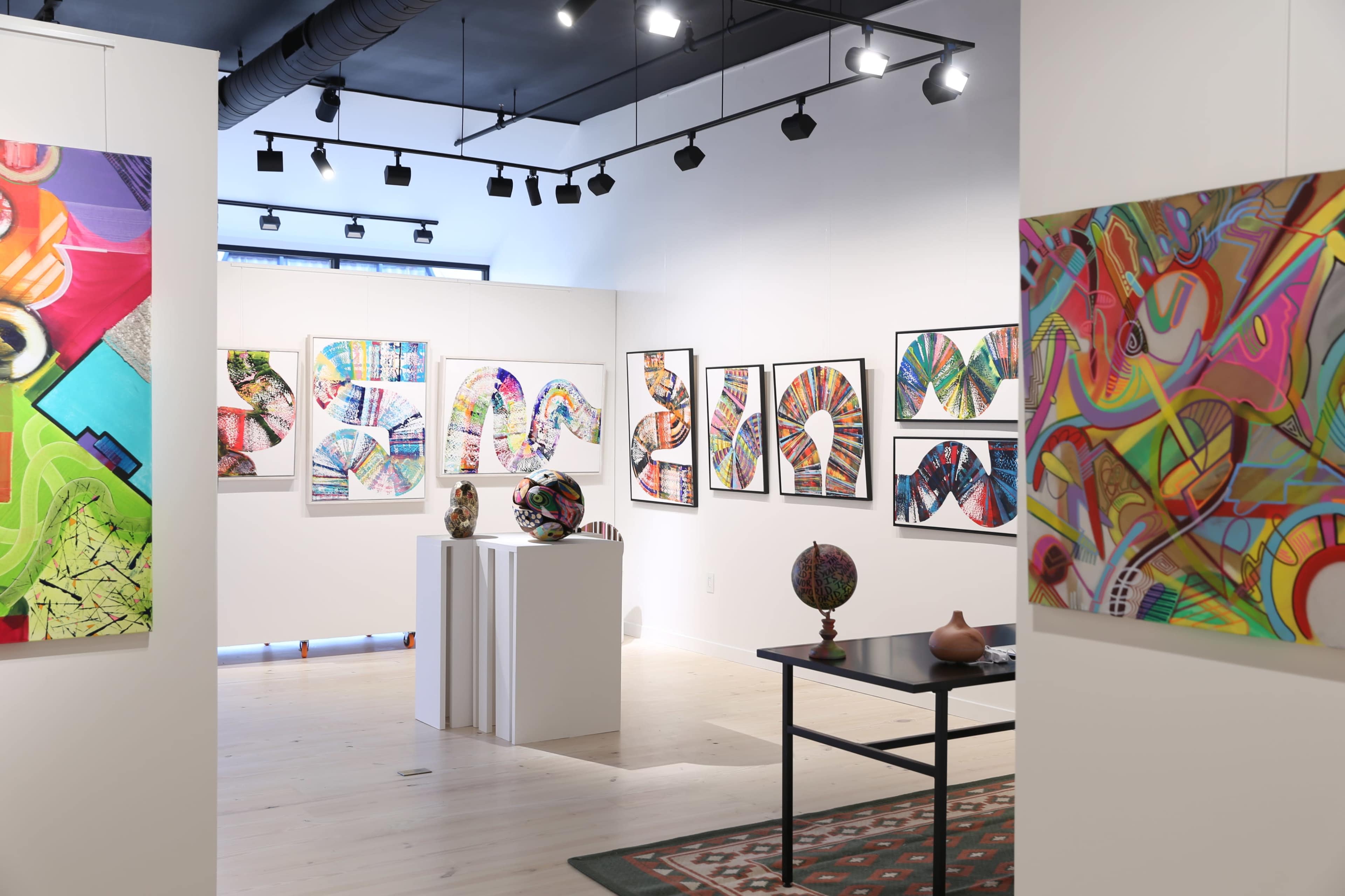An art gallery interior featuring colorful abstract artworks displayed on the walls and various sculptures on pedestals and tables.