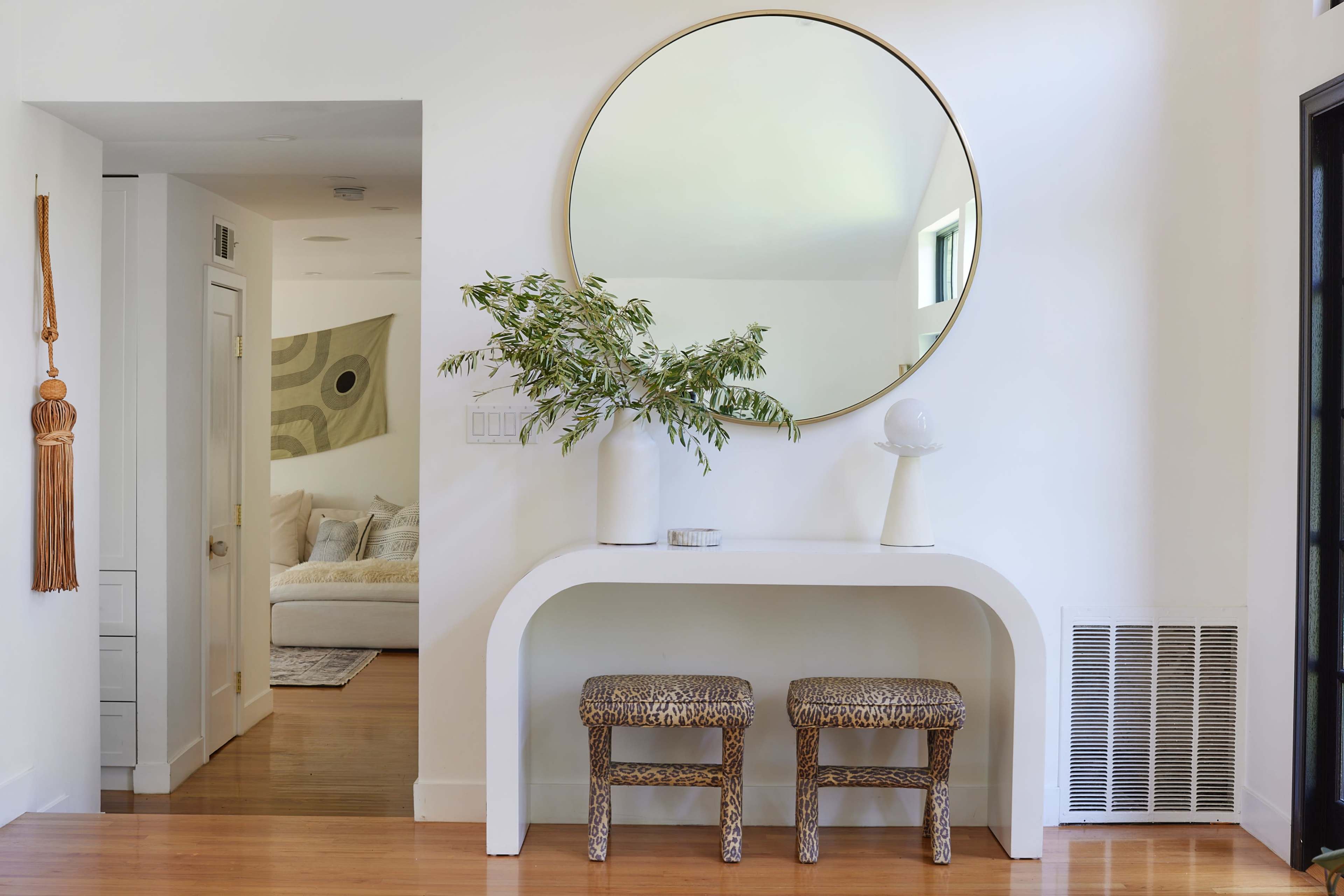 A minimalist entryway features a round mirror above a white console table flanked by two leopard print stools and a vase with greenery.