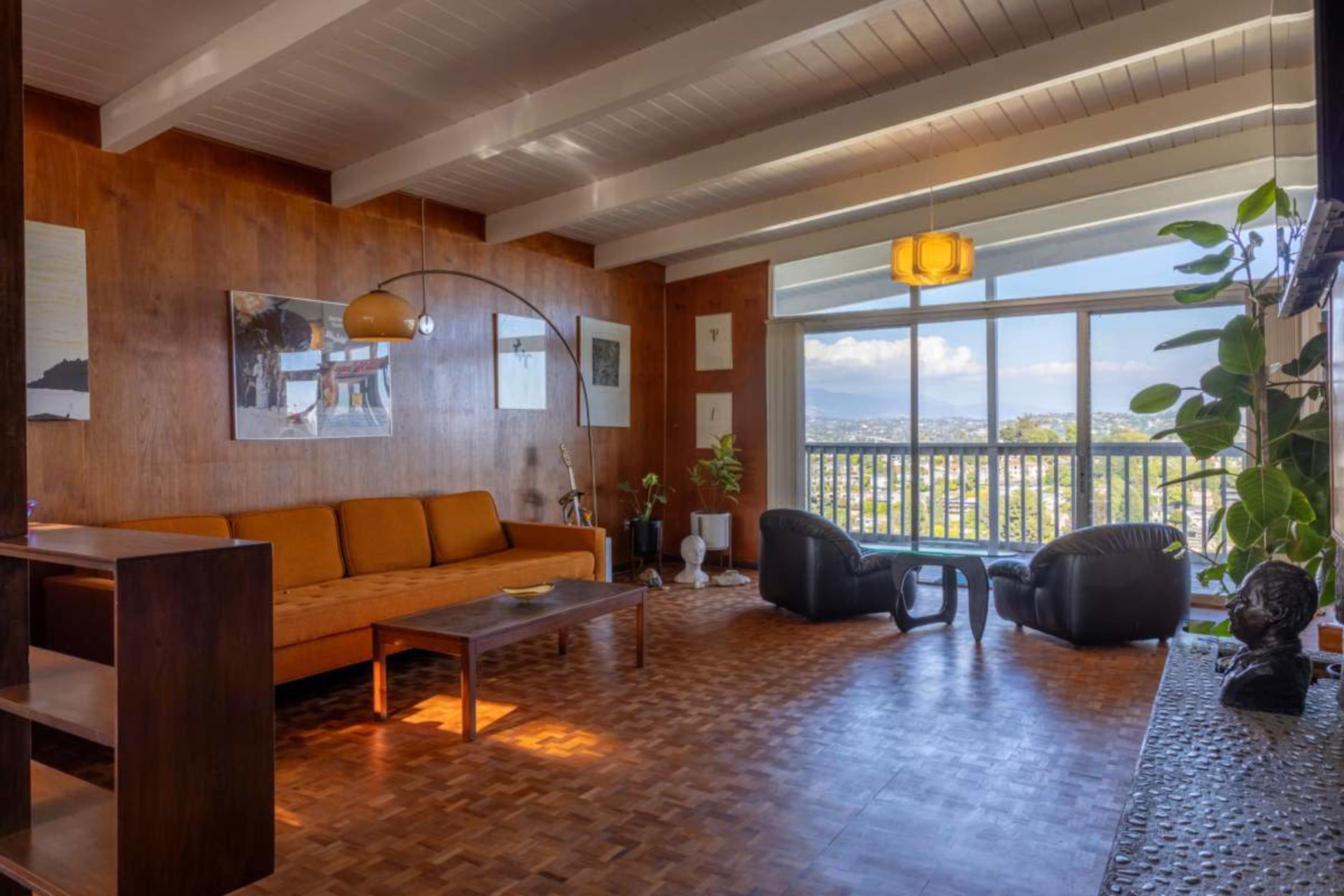 The image shows a spacious living room with a large orange sofa, a coffee table, and floor-to-ceiling windows that offer a view of the outdoors.