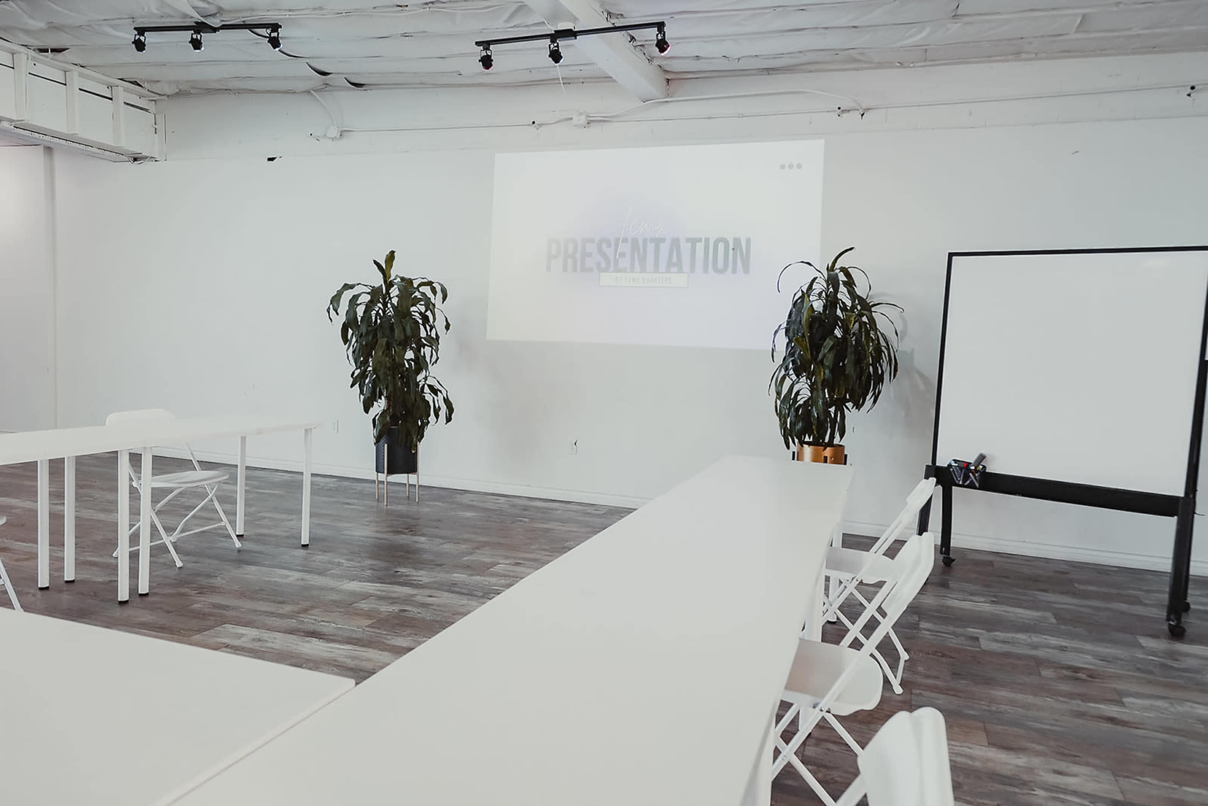 A bright, minimalist meeting room featuring white tables and chairs, with a projected presentation on a wall and potted plants on either side.