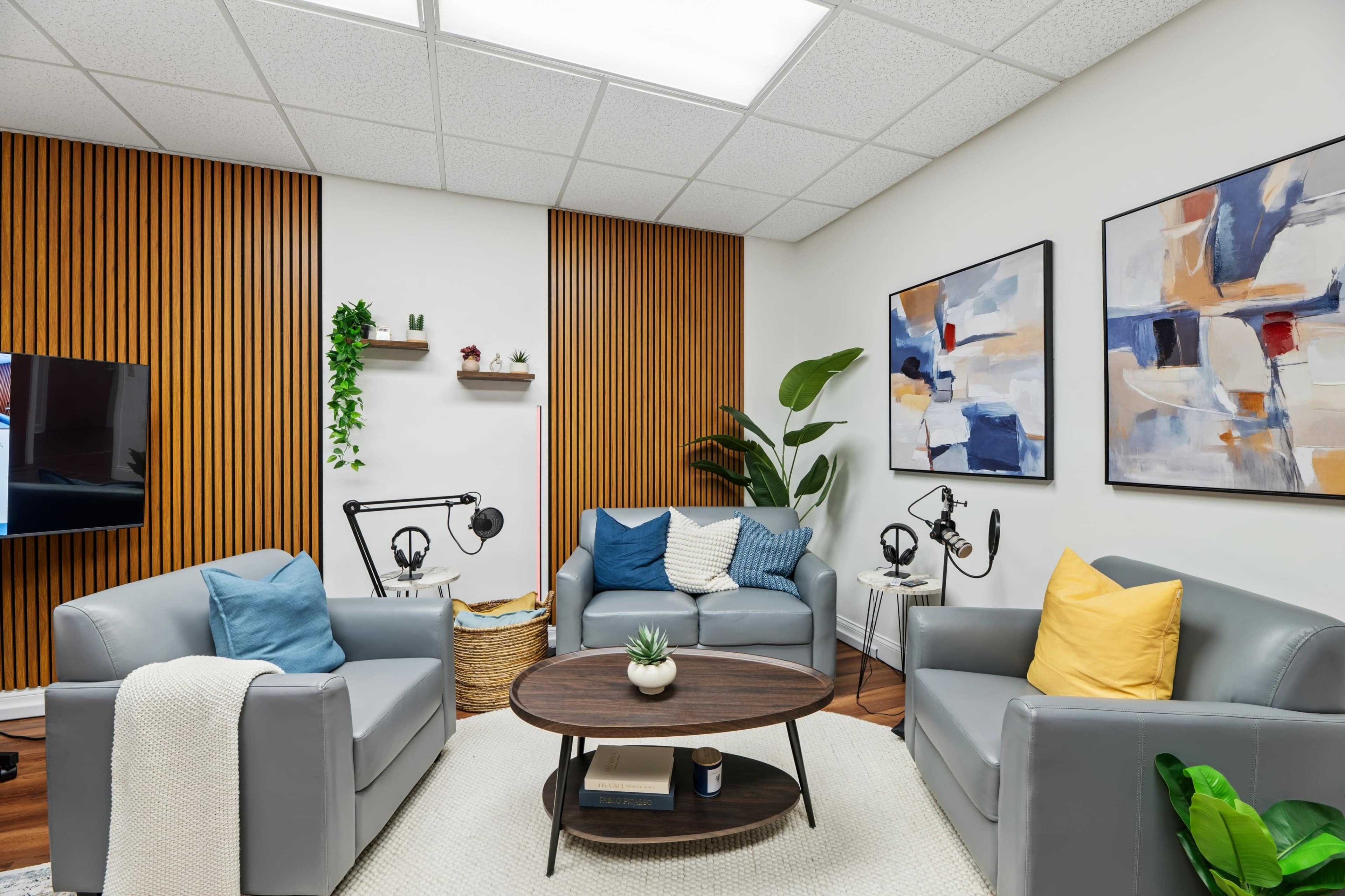 The image shows a modern, cozy sitting area with two gray sofas, a round wooden coffee table, and decorative wall art, set against wooden panel accents.